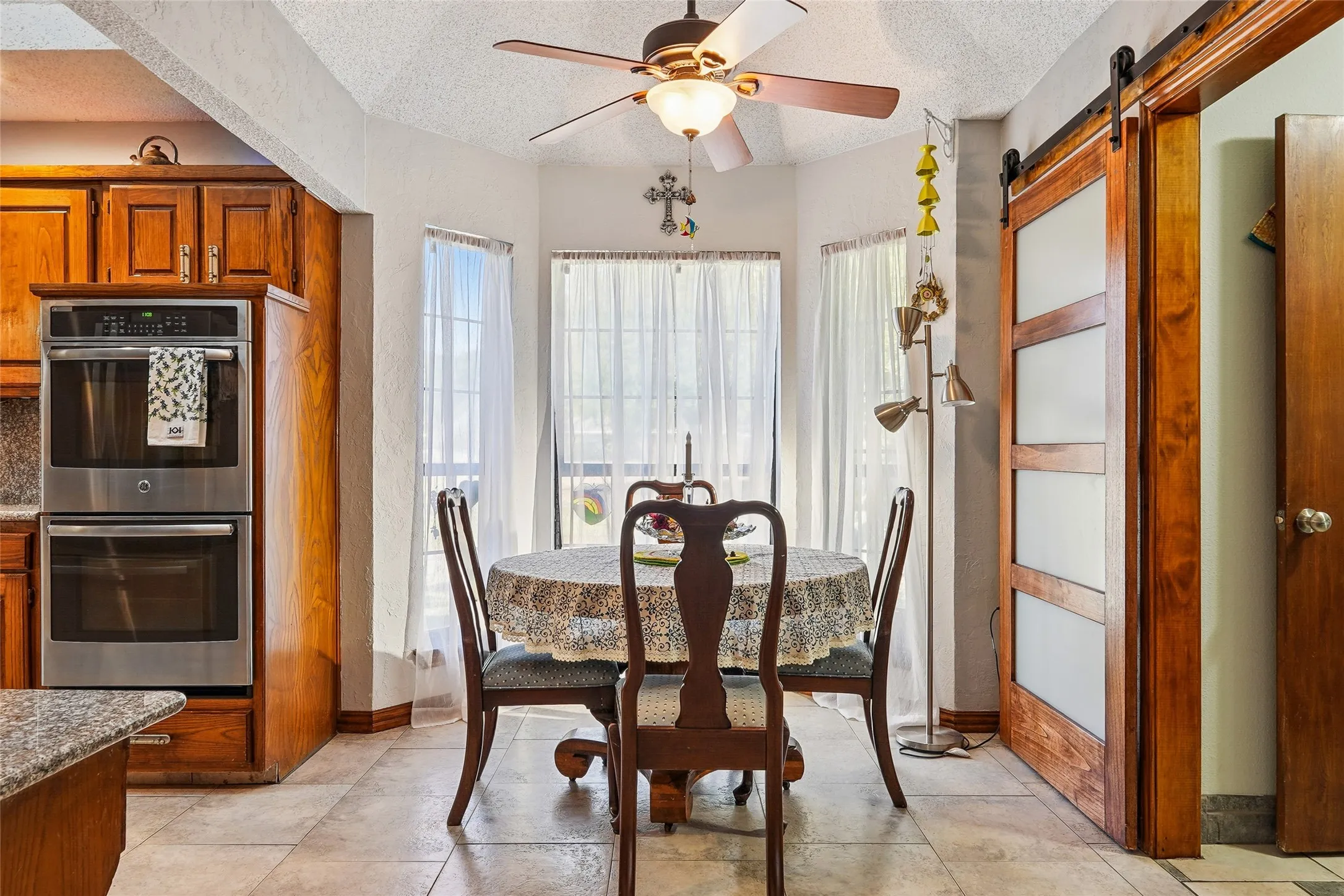 Bay window with multiple panels, ceiling fan with integrated lighting, wood-finish cabinetry, stainless steel double oven, and a wood-paneled barn door