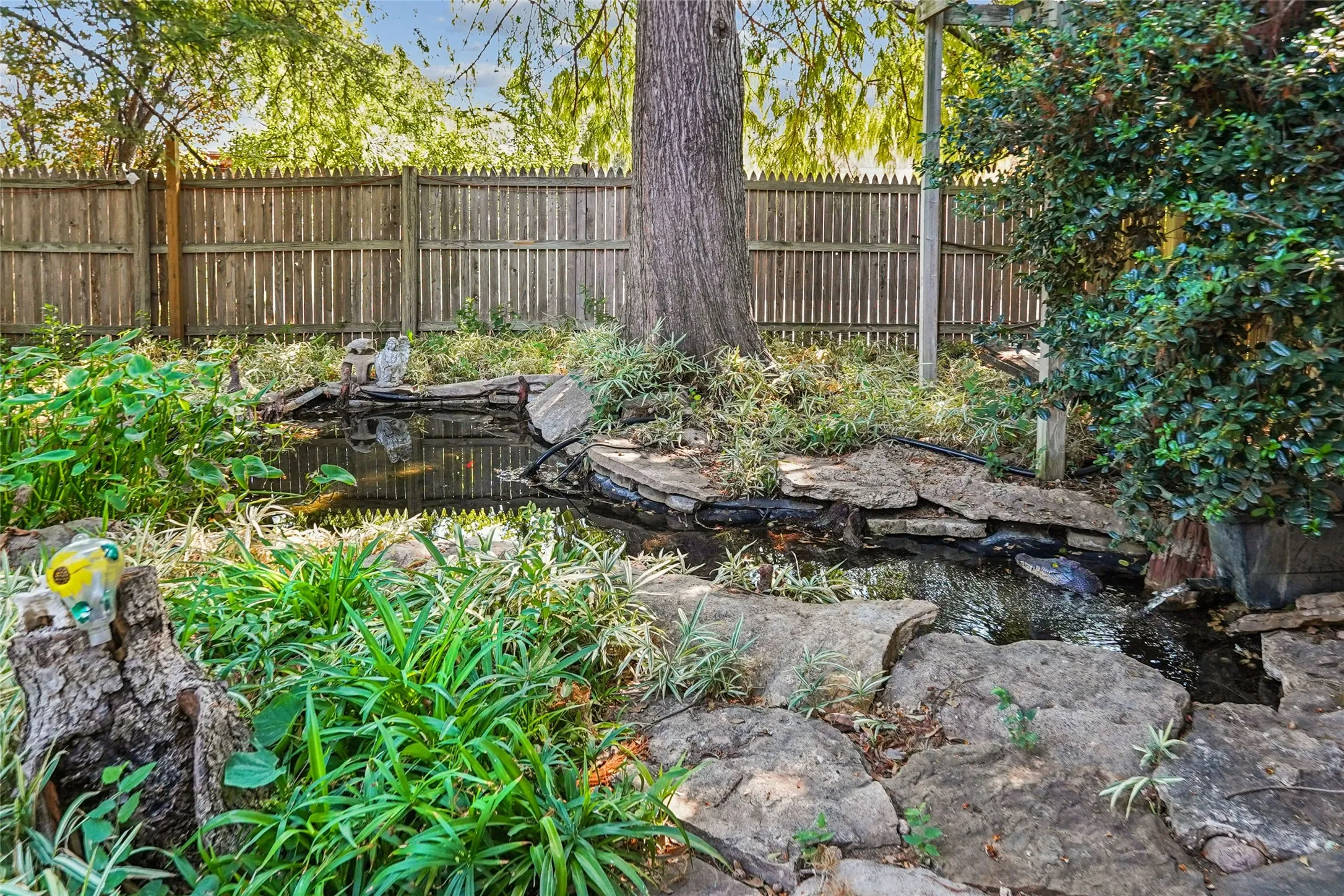 Landscaped yard featuring a water feature with stone edging, mature foliage, and a wood fence perimeter