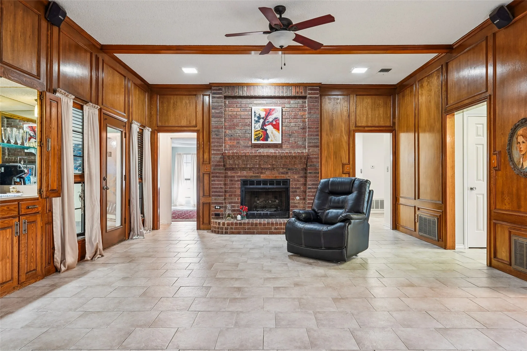 Full-height wood paneling with architectural trim, brick fireplace with a raised hearth, light-toned tiled flooring, and a ceiling fan with integrated lighting