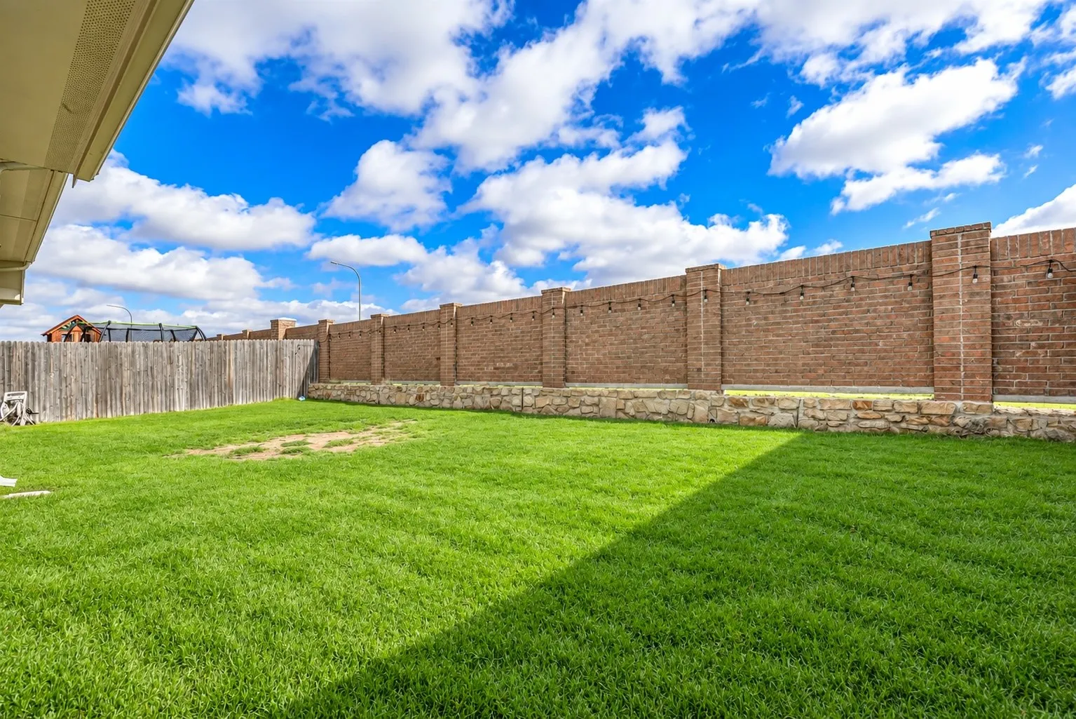 Expansive backyard featuring a manicured lawn, a full-height brick privacy fence with decorative string lighting, and a stone retaining wall