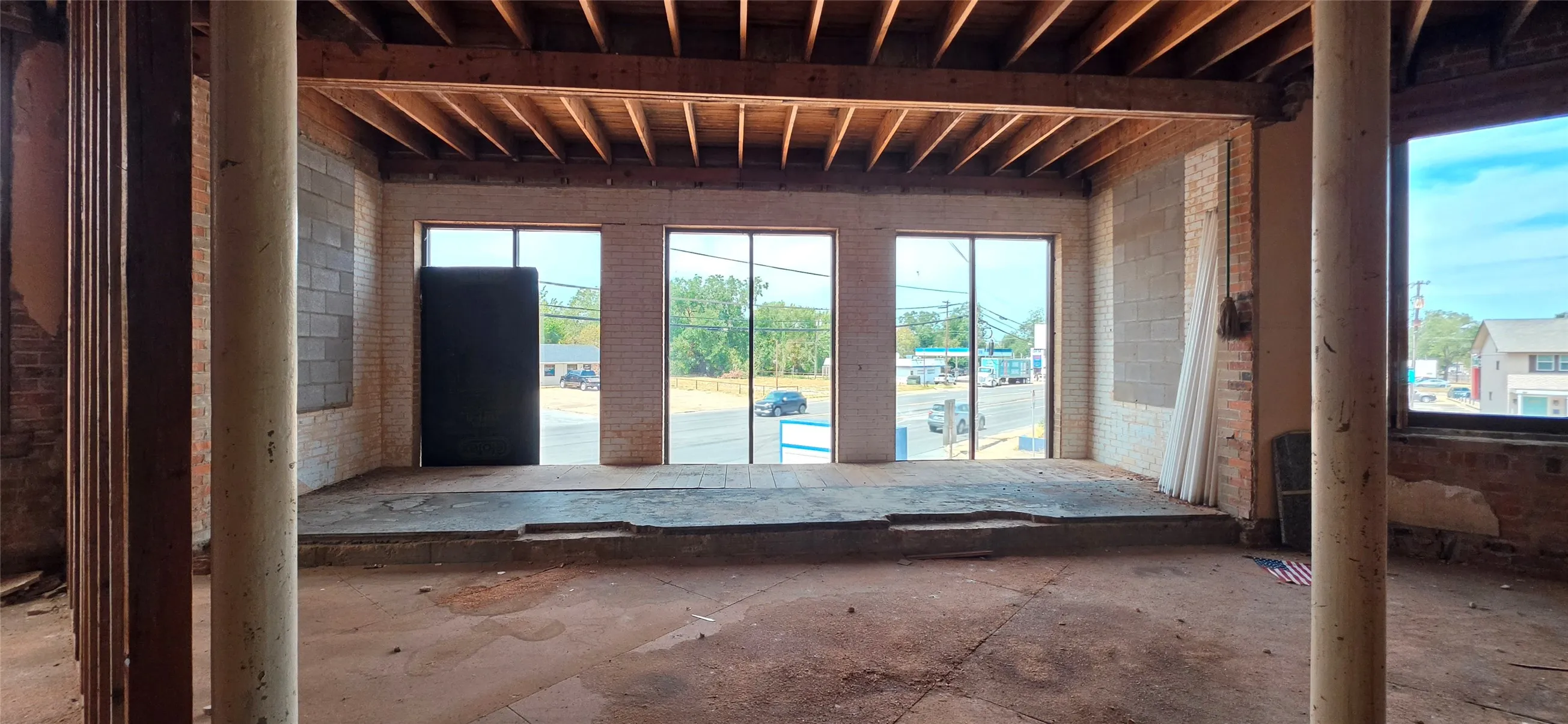 Exposed wood ceiling beams, three large window panels, bare brick wall, bare concrete floor, and structural support columns