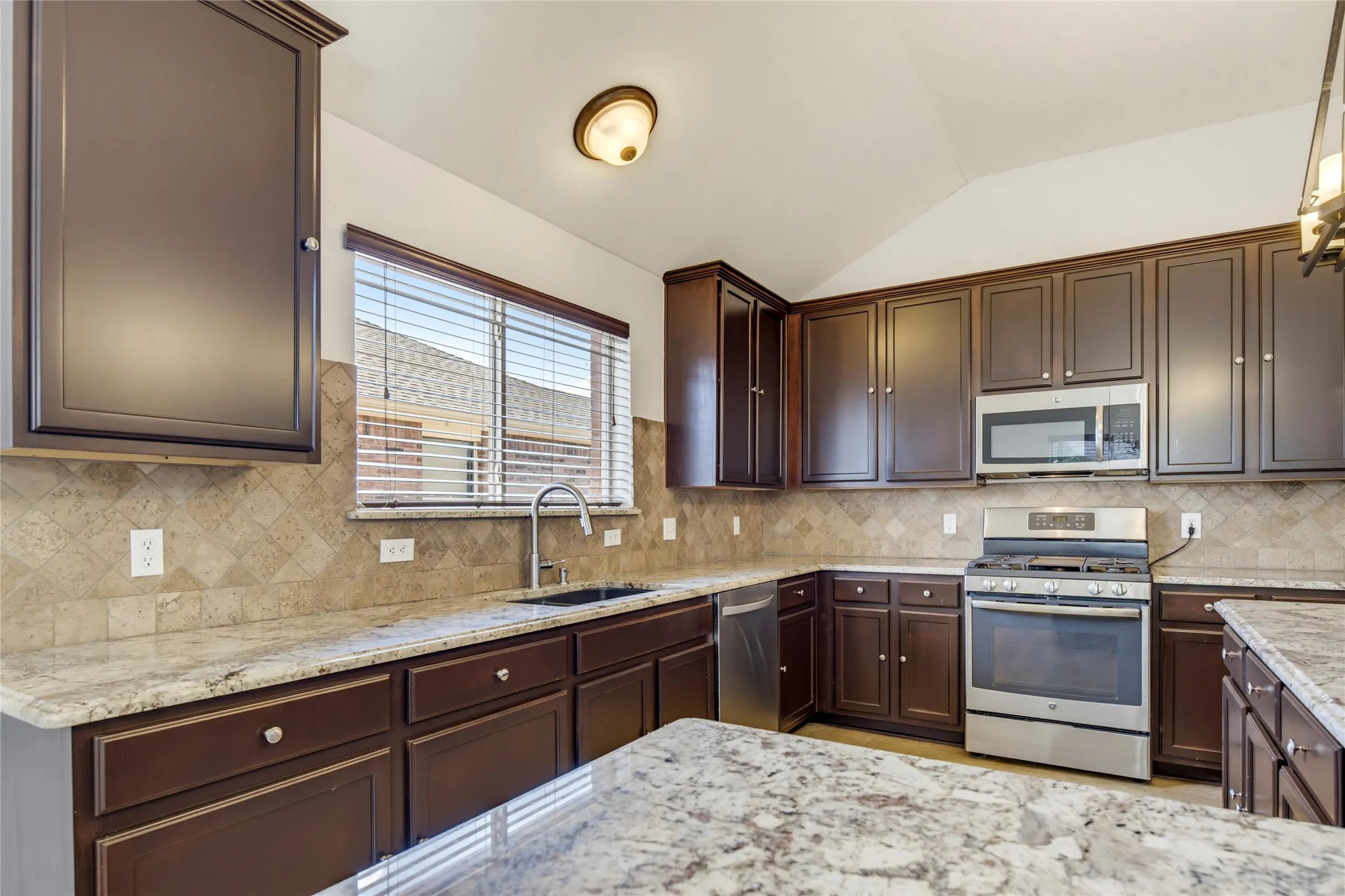 Kitchen featuring dark wood-finish cabinetry, light granite countertops, stainless steel appliances, a tile backsplash, and a vaulted ceiling
