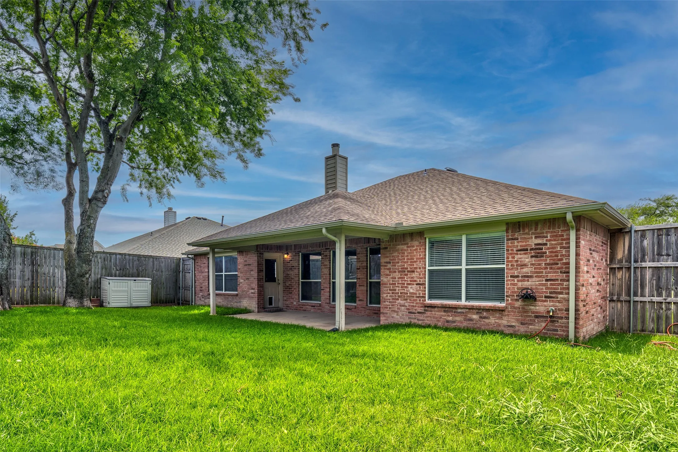 Expansive green lawn with mature shade tree, brick exterior, covered patio, and privacy fencing