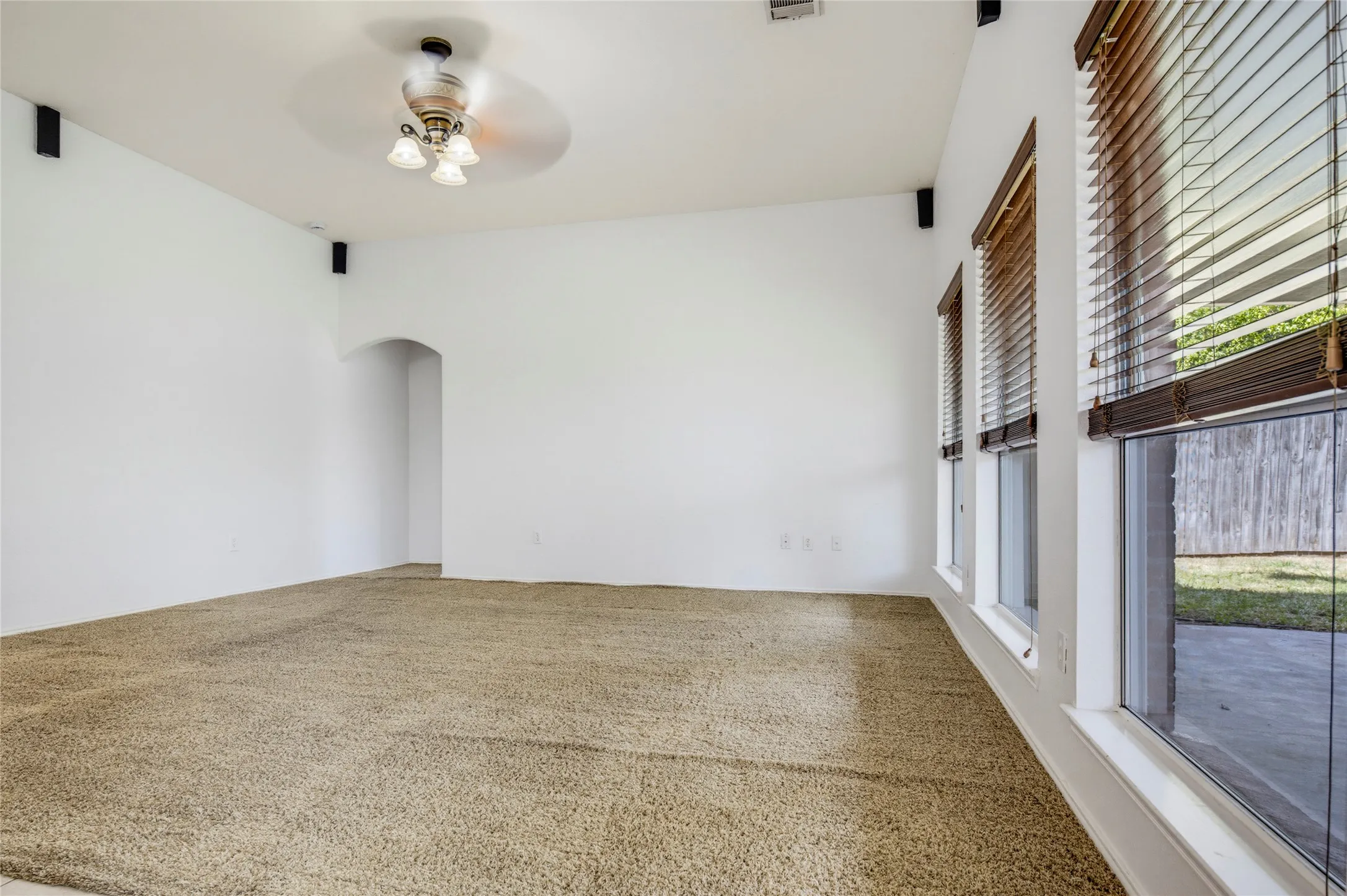 Textured carpet flooring, multiple windows with wood-finish blinds, arched doorway, ceiling fan with light fixture, built-in speakers