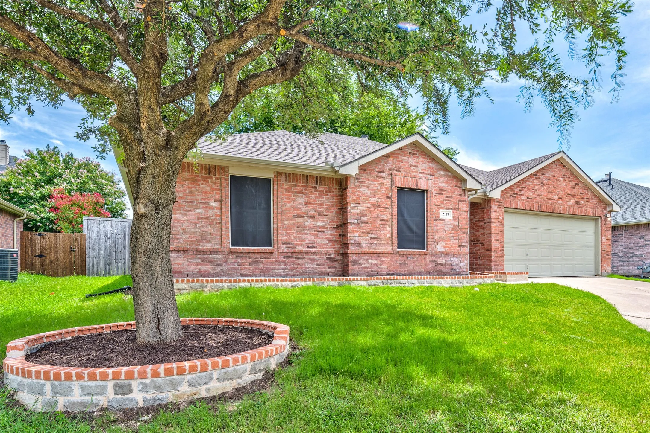 Traditional brick exterior with an attached garage, a mature tree with a stone and brick planter, a shingled roof, and a concrete driveway