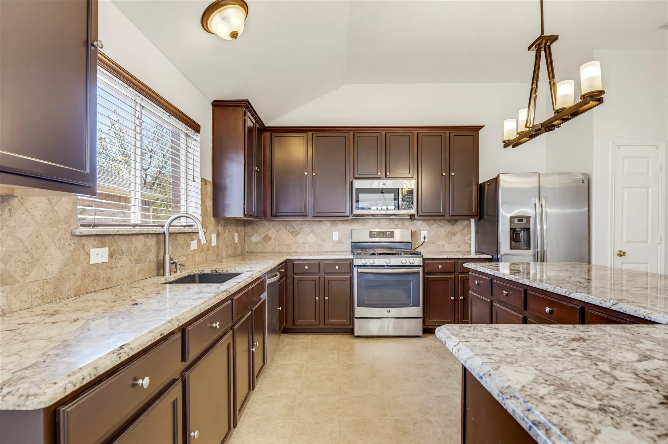 Kitchen featuring dark wood cabinetry, light-toned countertops, stainless steel appliances, a tiled backsplash, and vaulted ceilings