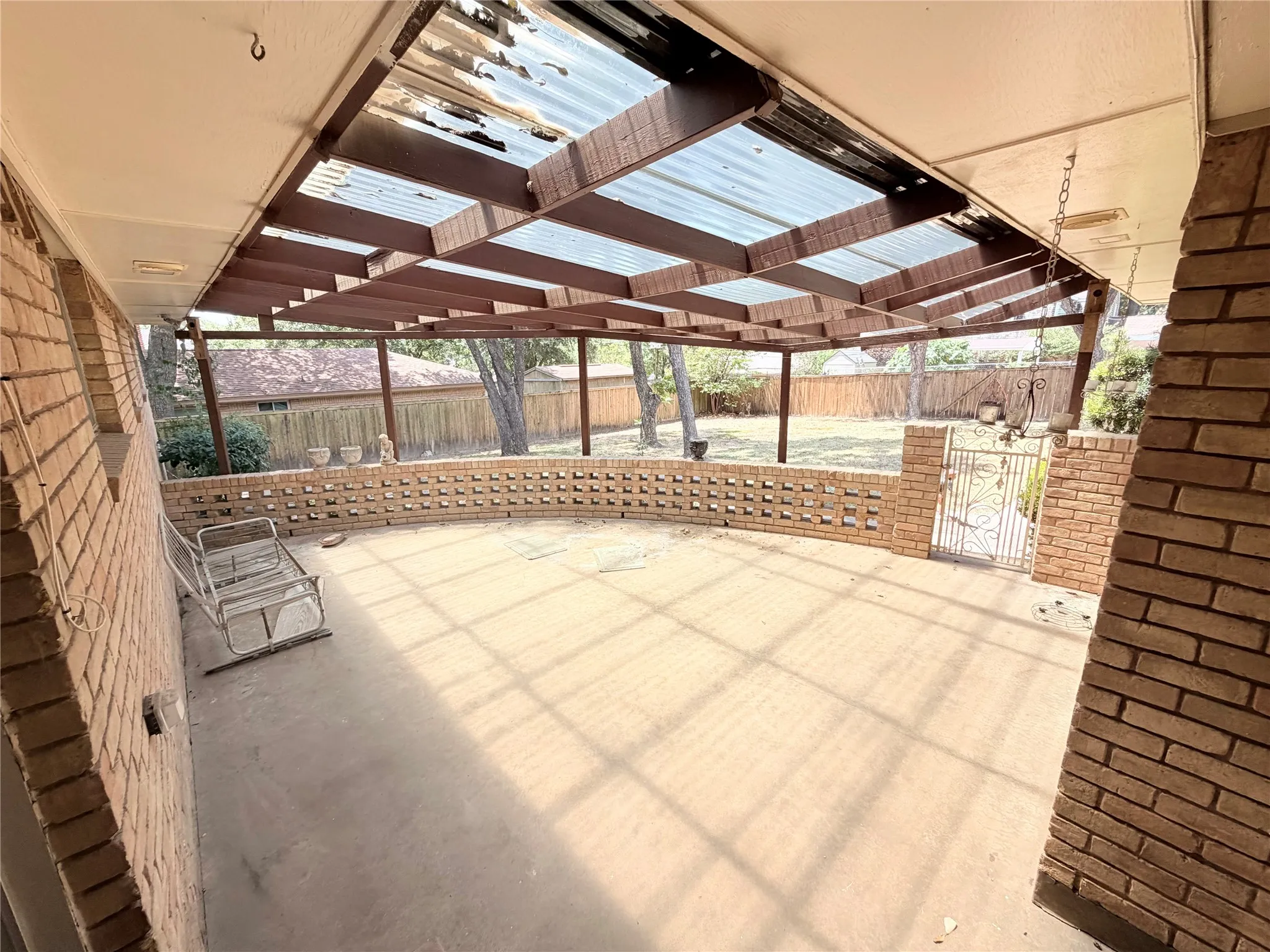 Expansive covered patio with a concrete surface, brick perimeter wall, and a decorative metal gate, all under a wood-beamed translucent roof structure