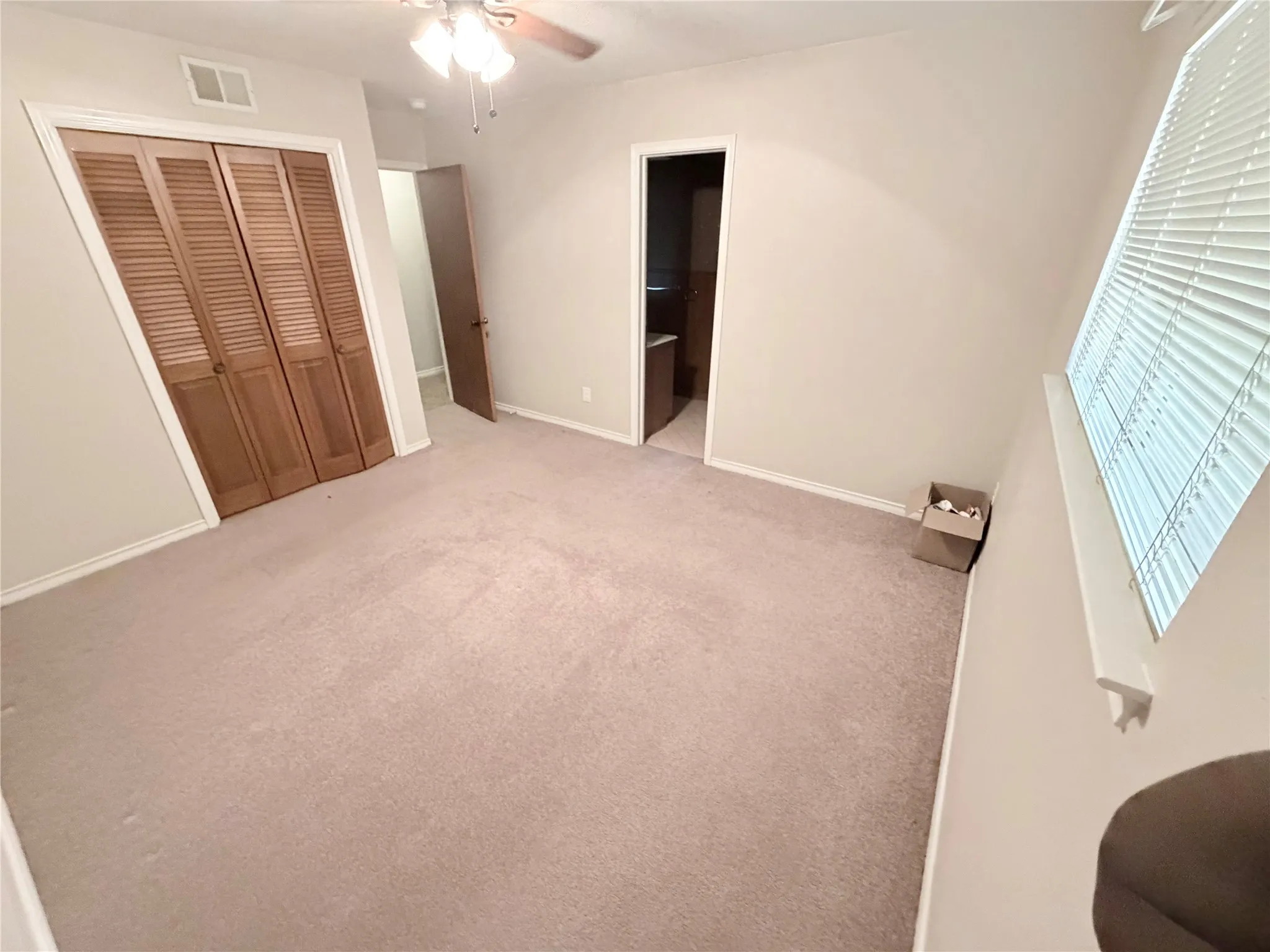 Neutral tone wall paint, carpeted flooring, window with horizontal blinds, bi-fold louvered closet doors, and a ceiling fan with light fixture