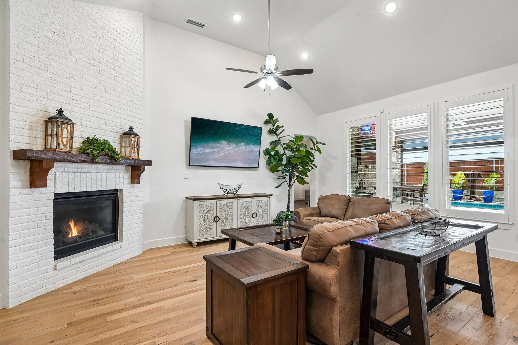 Vaulted ceiling with recessed lighting, whitewashed brick fireplace with wood mantel, wood-finish flooring, ceiling fan, and plantation shutters