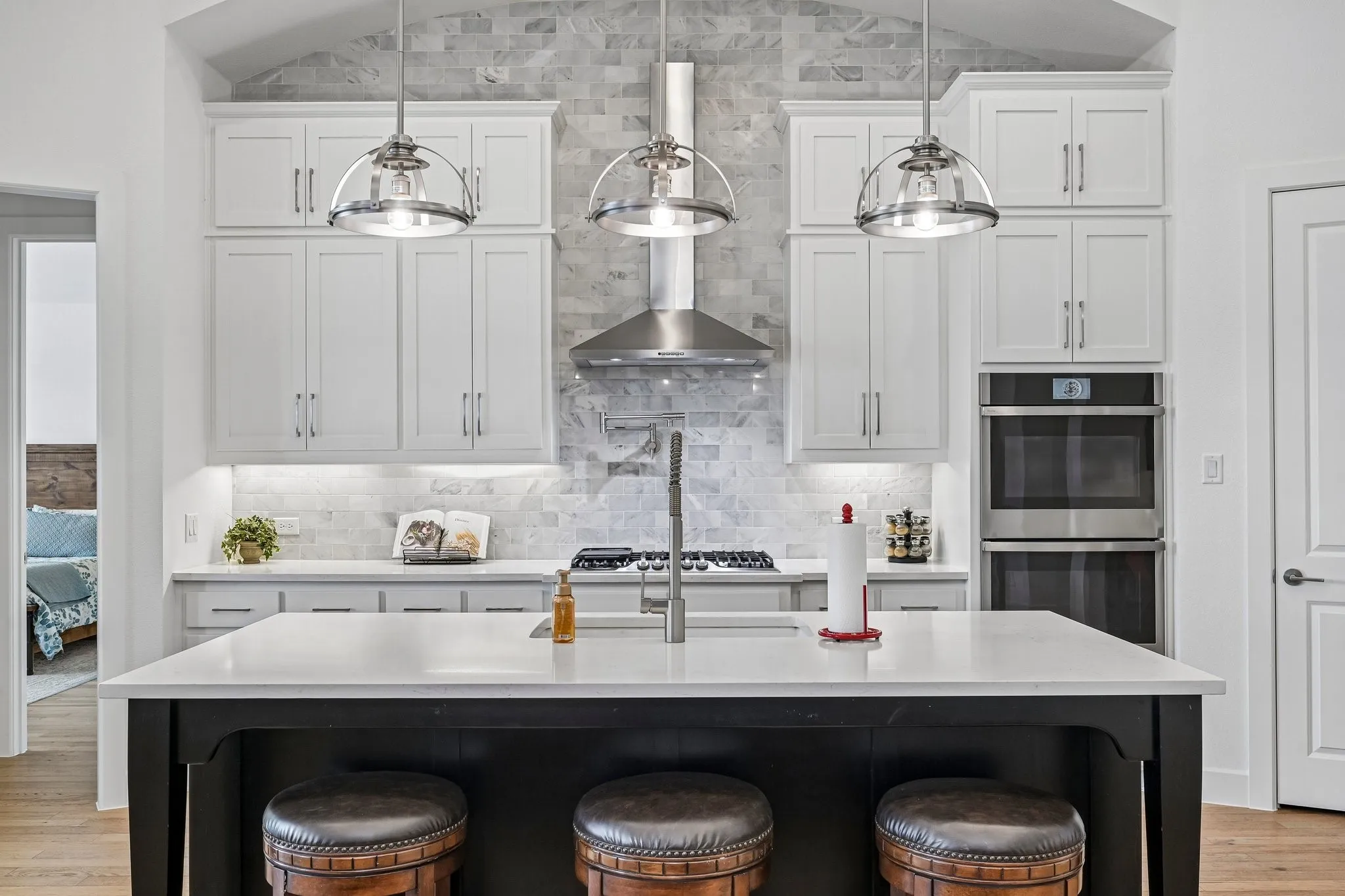 White shaker-style cabinetry, stainless steel appliances, stone backsplash, large center island with counter seating, and three pendant light fixtures