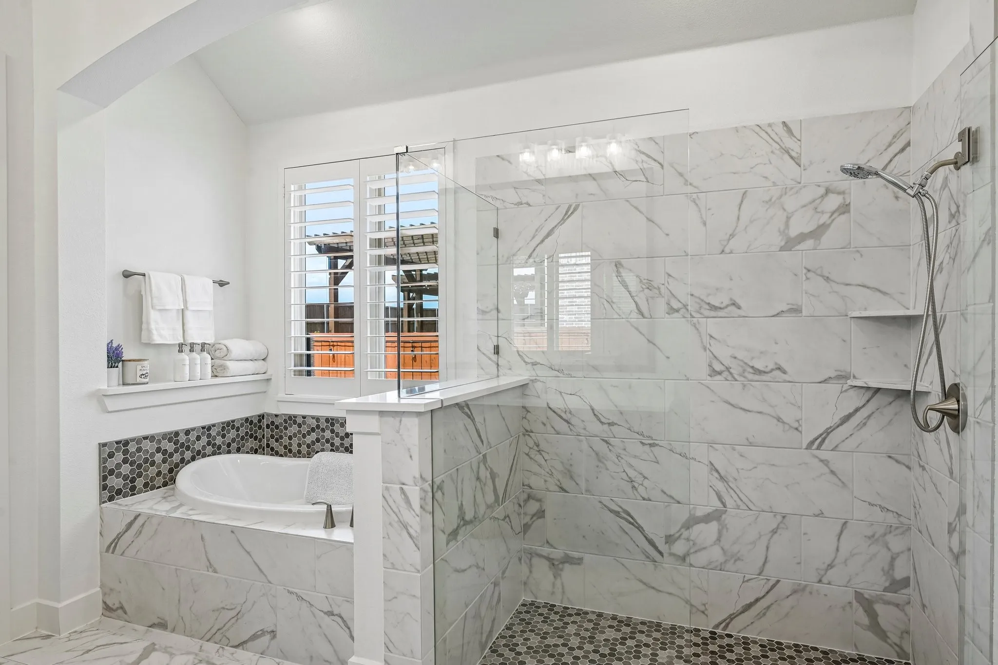 Bathtub with tile surround, walk-in shower with glass enclosure, mosaic tile shower floor, wall-mounted showerhead, and window with plantation shutters