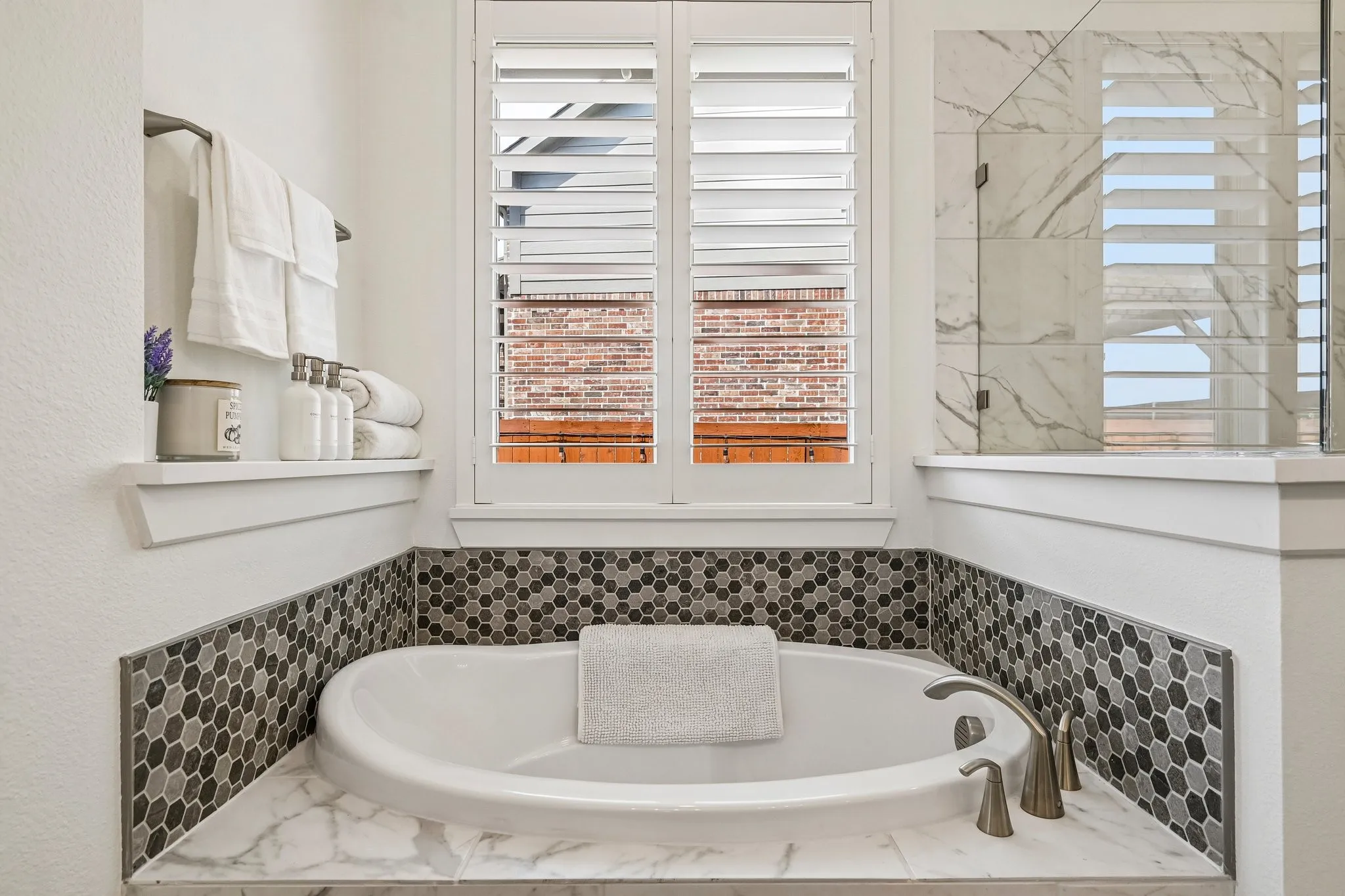 Built-in soaking tub with a brushed nickel faucet, hexagonal mosaic tile surround, and plantation shutters