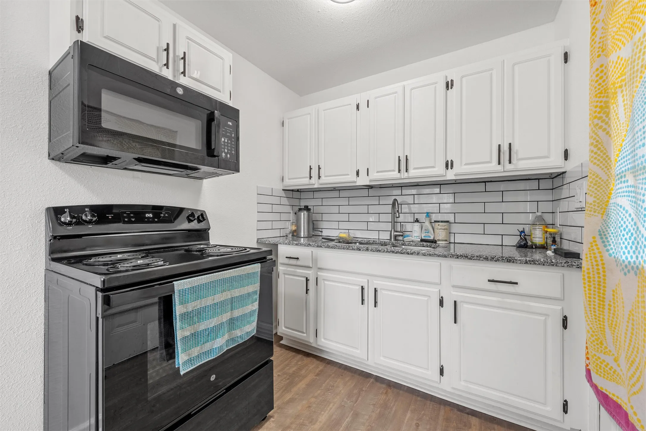 White shaker-style cabinetry with matte black hardware, subway tile backsplash, granite-style countertops, stainless steel sink with a high-arc faucet, and wood-finish flooring