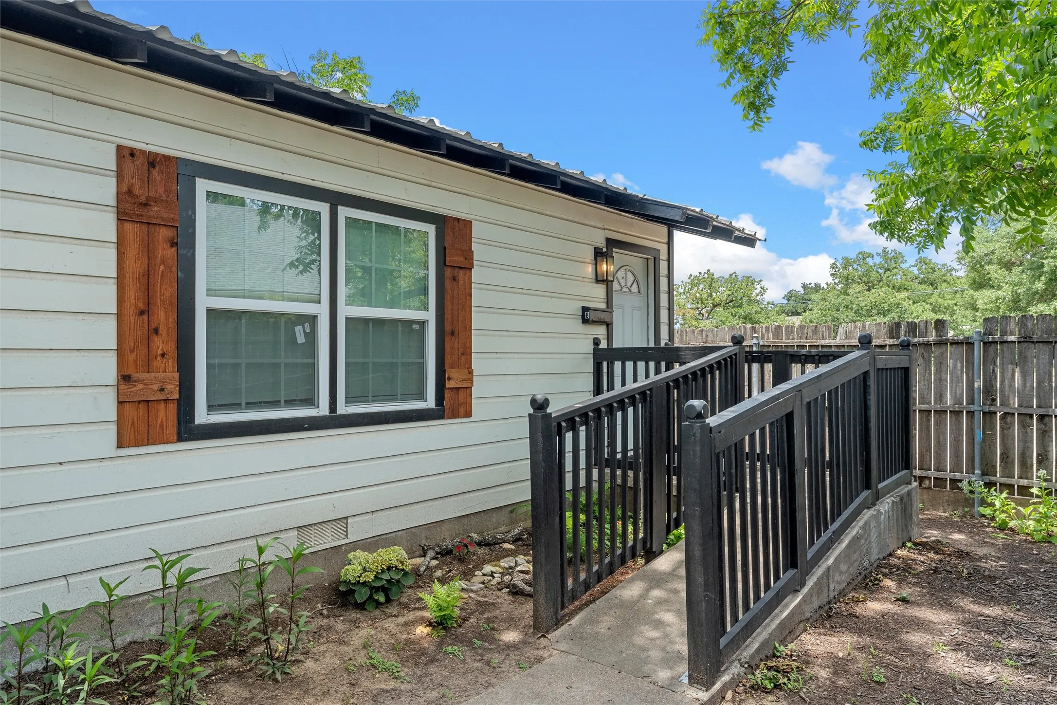 White siding exterior with a black trim window, wood shutters, black metal railing, and a wood privacy fence