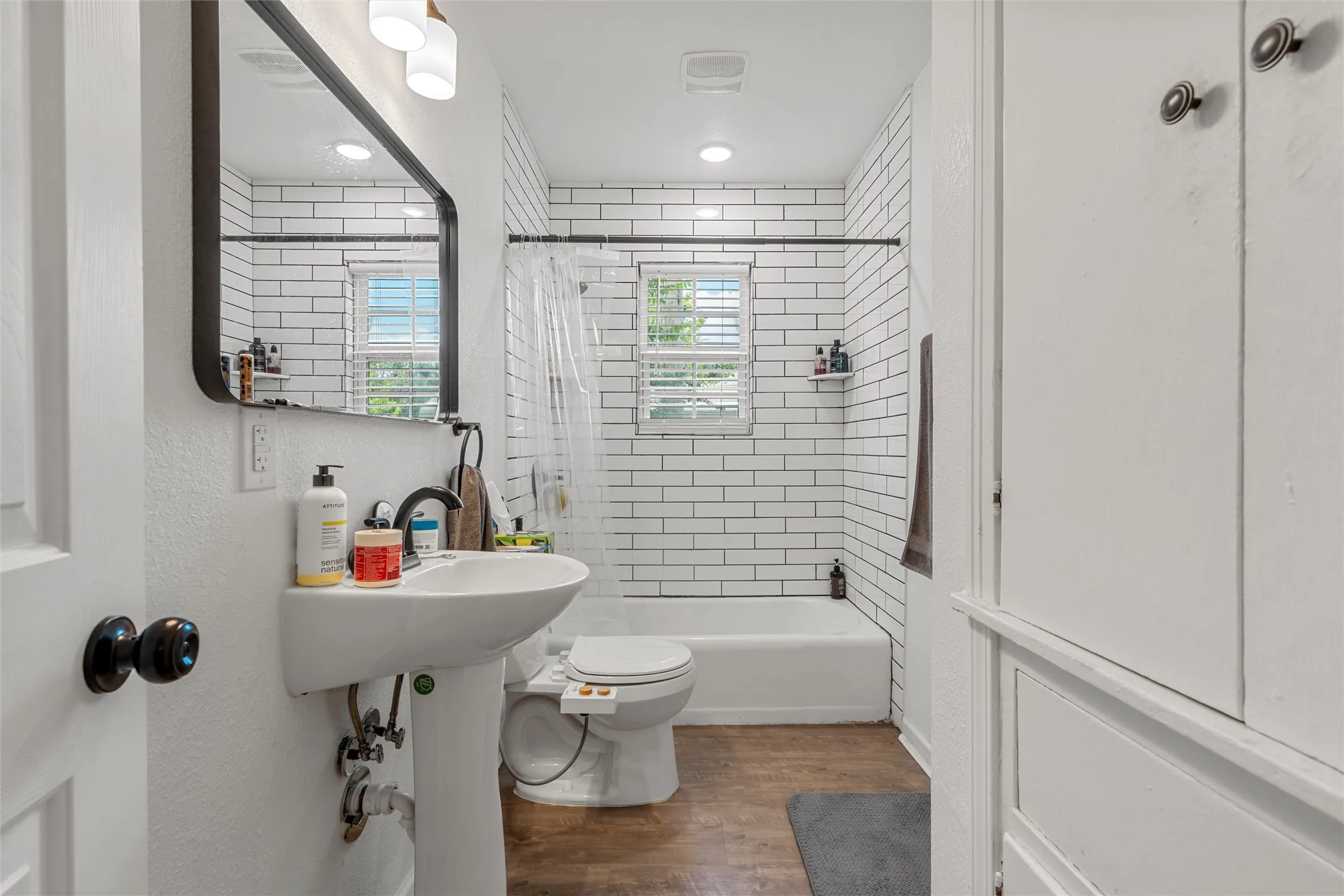 Pedestal sink with dark-toned fixtures, rectangular wall mirror with a dark frame, white subway tile shower surround, window with blinds, and built-in storage cabinetry