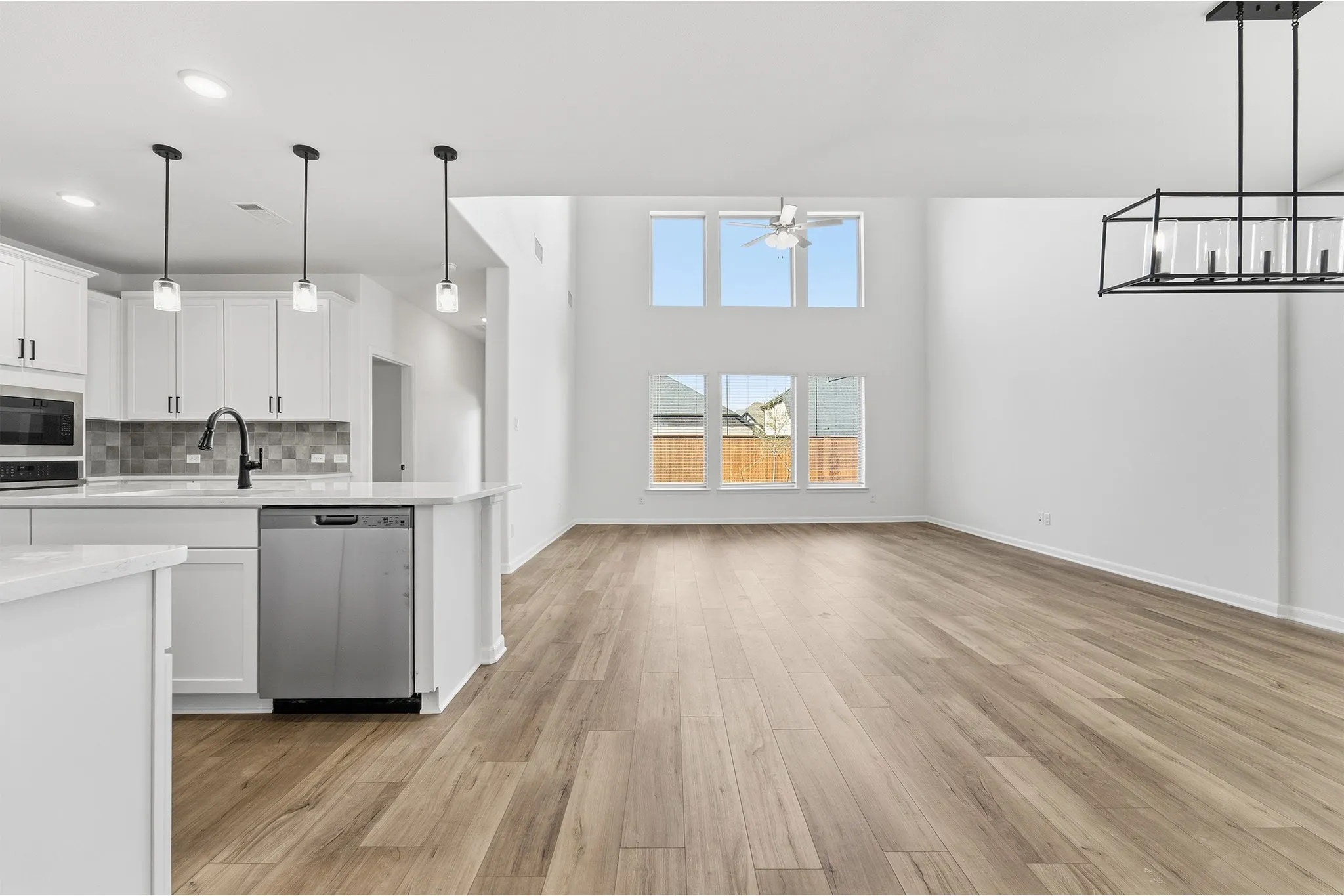 Open-concept layout featuring wood-finish flooring, a kitchen island with pendant lighting, stainless steel appliances, and a two-story living area with high windows and a contemporary chandelier