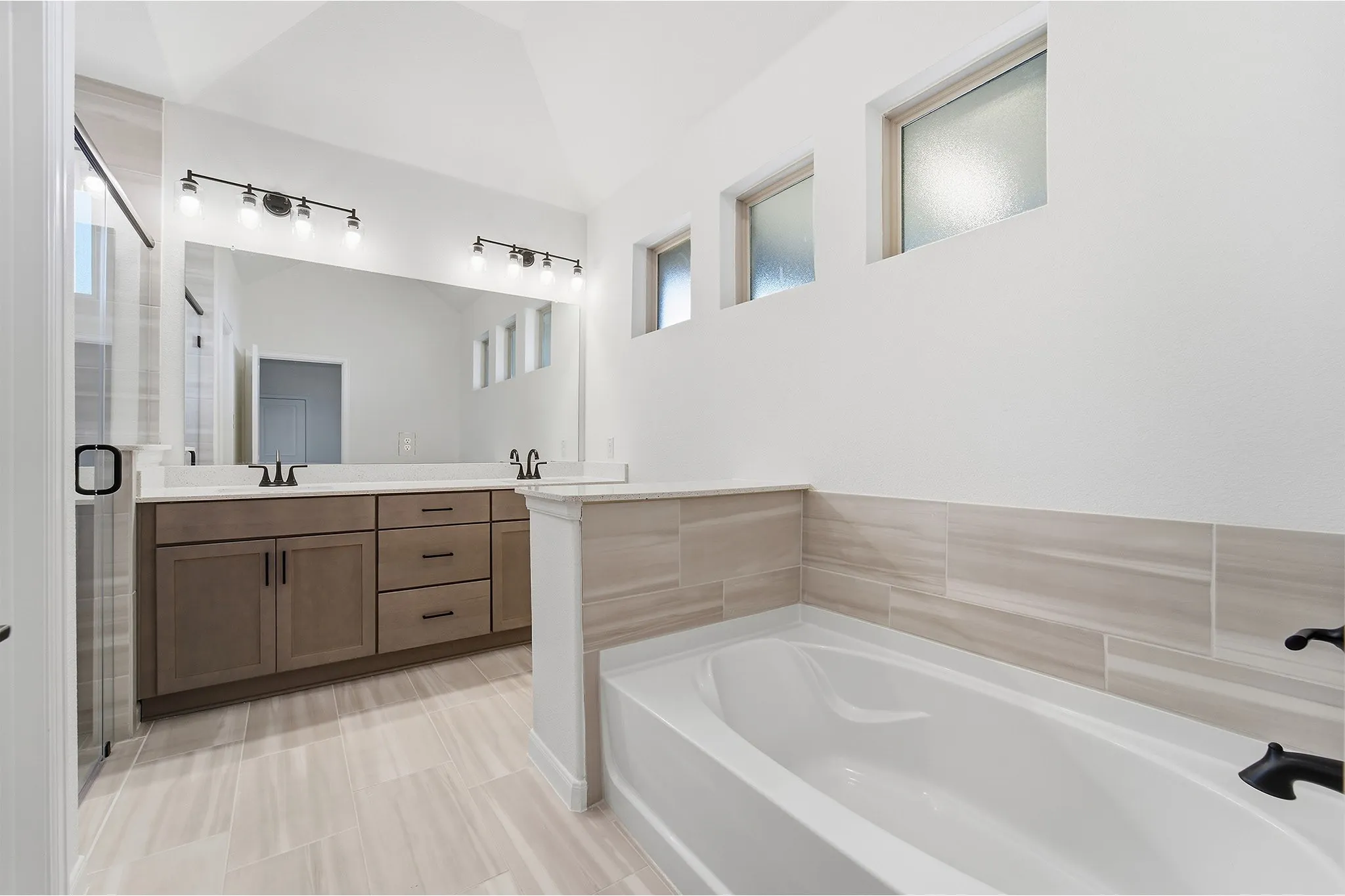 Dual vanity with stone countertops and undermount sinks, separate soaking tub, walk-in shower with glass enclosure, large format tile flooring, and multiple high windows