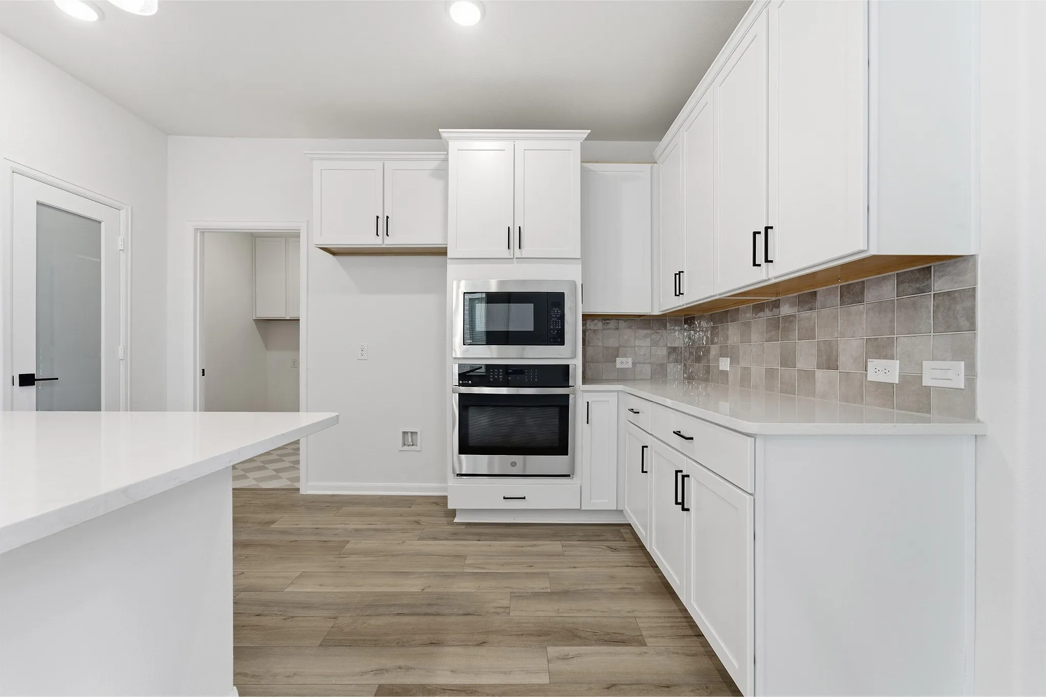 White shaker-style cabinetry, stainless steel built-in appliances, light-toned wood-finish flooring, and a tile backsplash