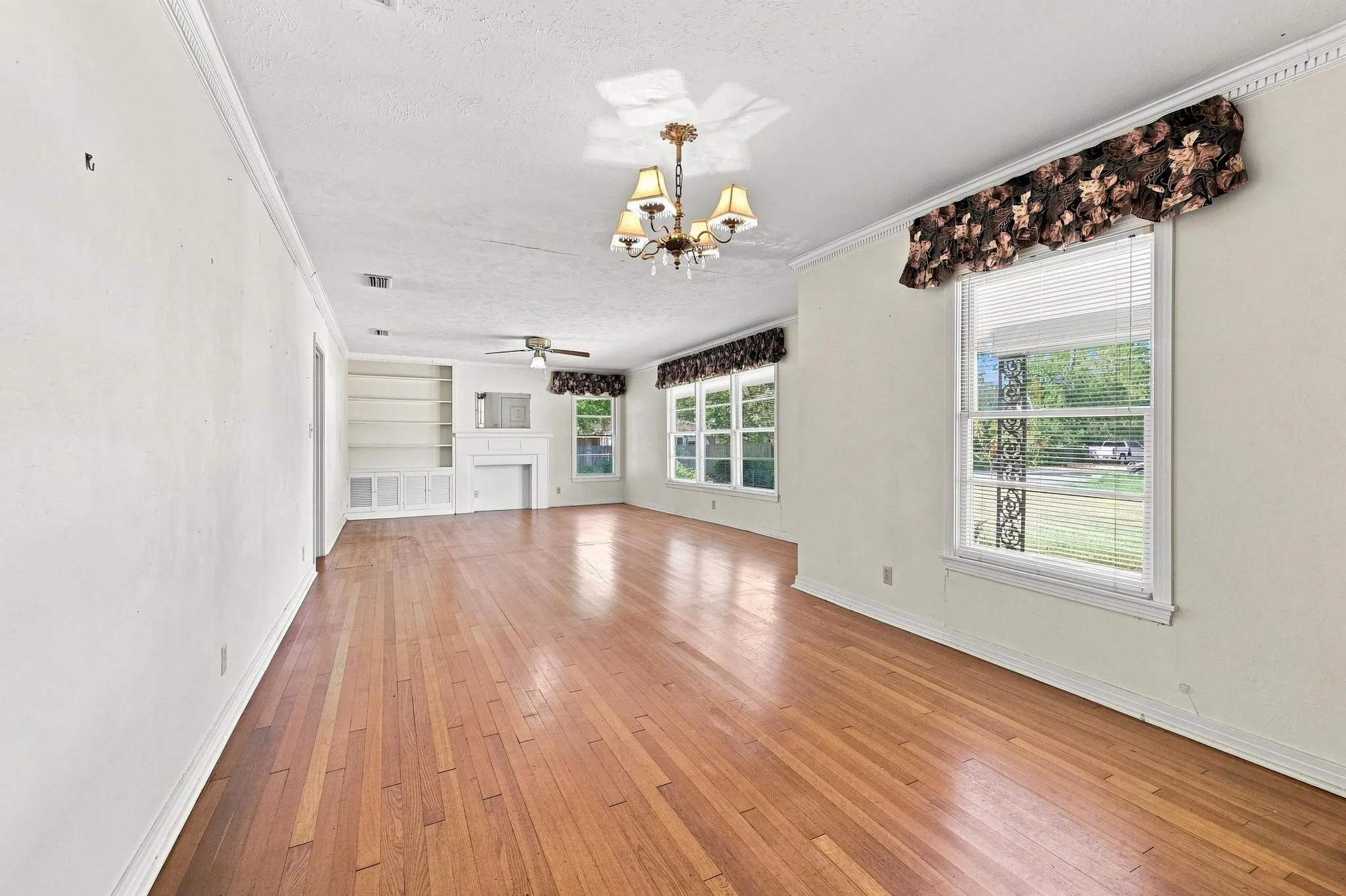 Hardwood flooring throughout this area, built-in shelving, faux fireplace inset, multiple windows, ceiling fan, and crown molding.