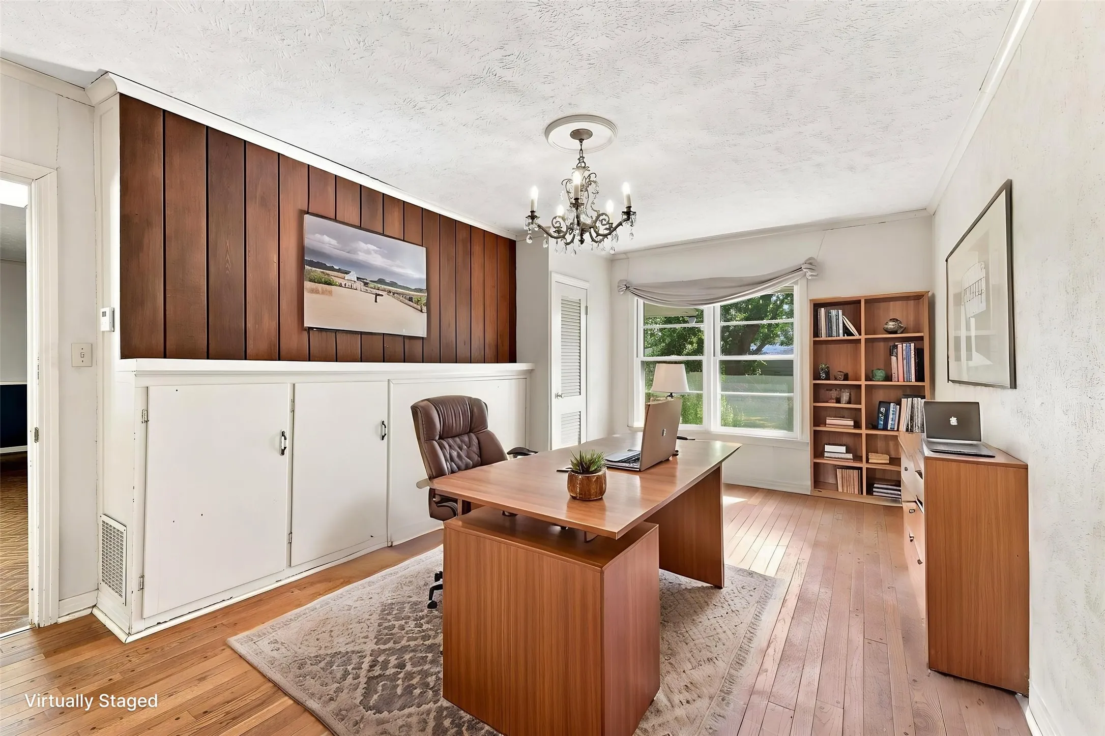 Hardwood flooring, wood panel accent wall, built-in lower cabinetry, large window, and chandelier.