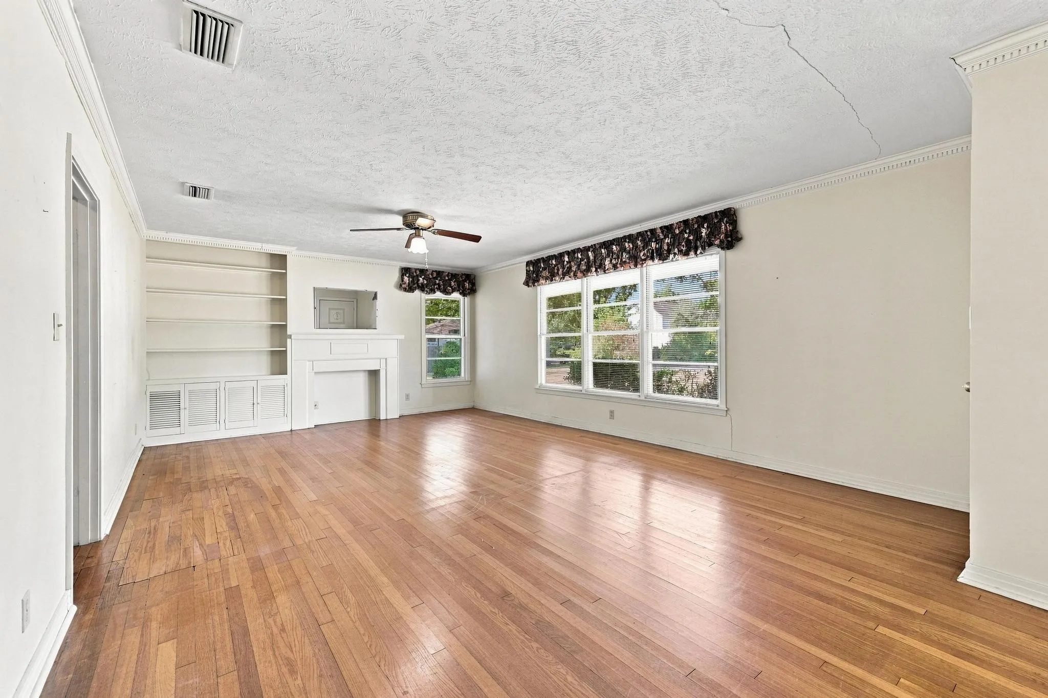 Hardwood flooring throughout, featuring a built-in shelving unit with lower cabinetry, a classic faux fireplace surround, and multiple large windows.