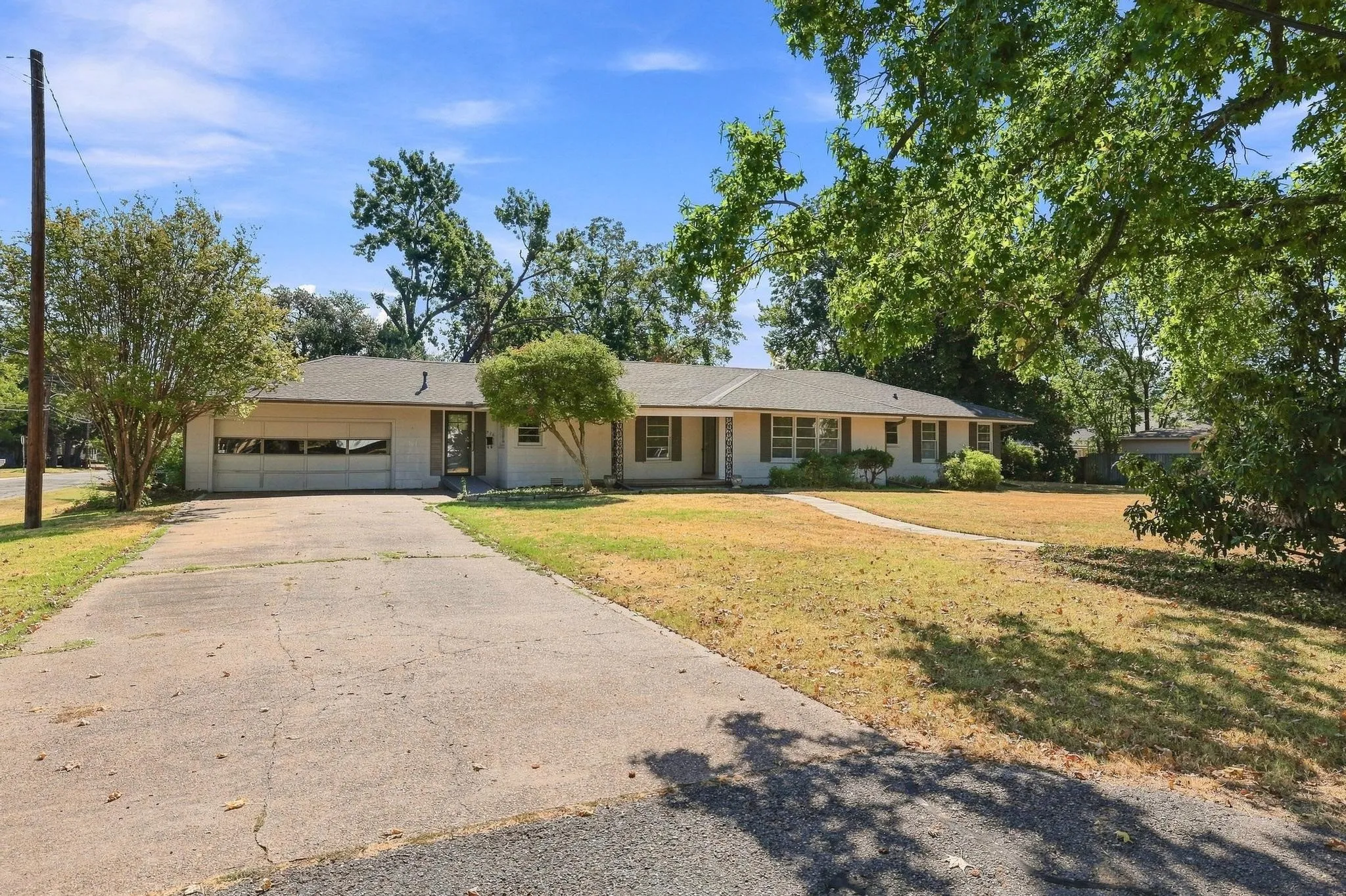 Single-story ranch-style featuring an attached garage, a paved driveway, and a front lawn with mature trees.