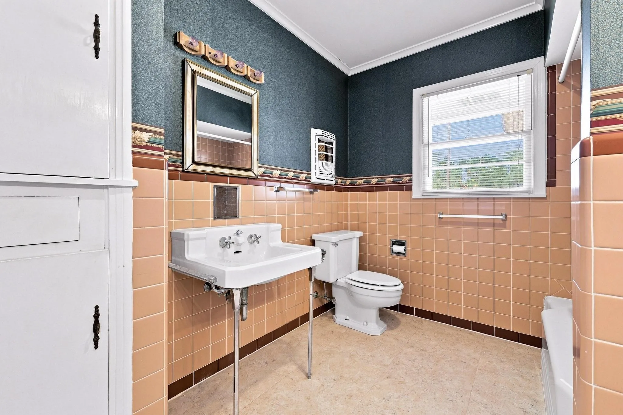 Built-in storage cabinet, pedestal sink, ceramic wall tiling, window with blinds, and a toilet. Vanity lighting can be turned on/off with a switch located on the right side of the light casing.