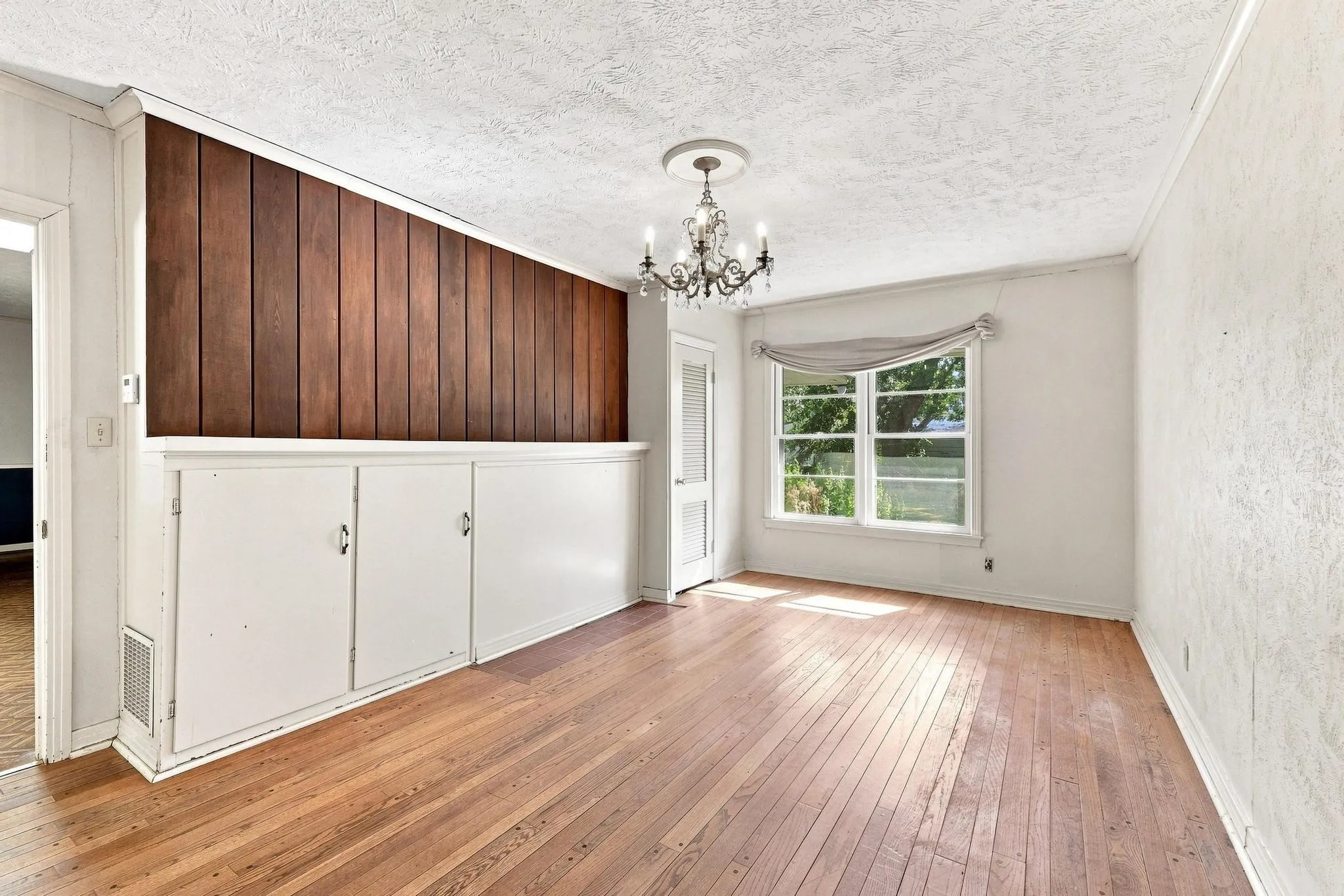 Hardwood flooring, built-in cabinetry, wood panel wall accent, window with valance, and decorative chandelier.