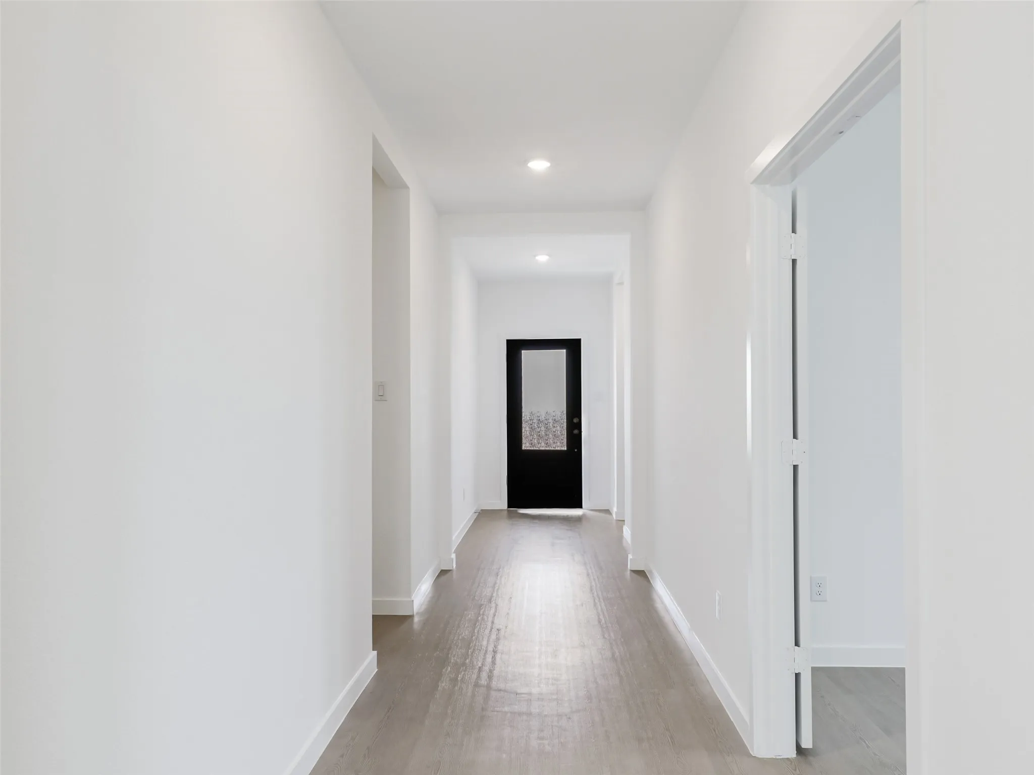 Recessed lighting illuminates the hallway, featuring light wood-finish flooring and a black door with frosted glass