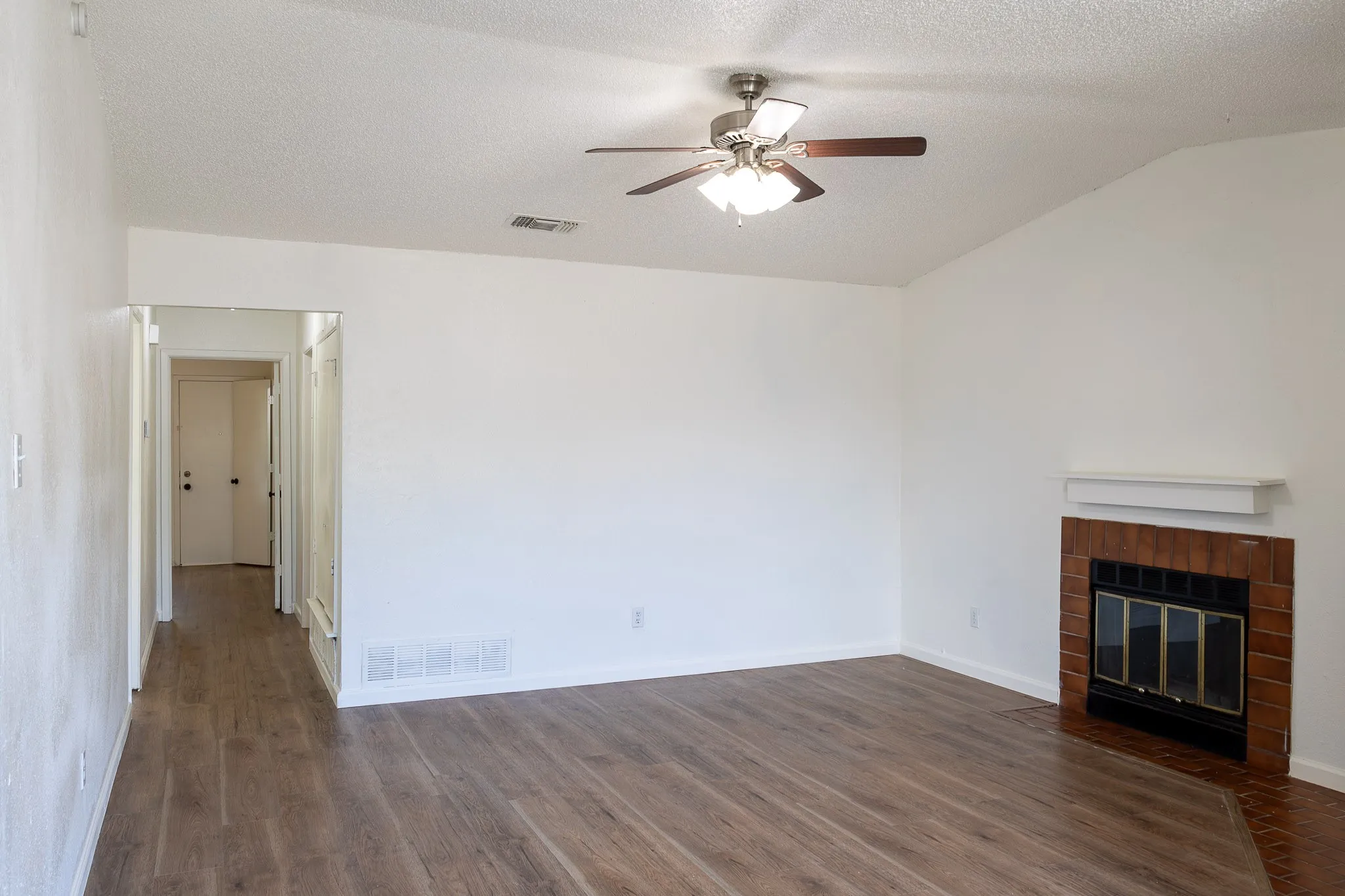 Vaulted ceiling, wood-finish flooring, brick fireplace with mantel, ceiling fan with integrated lighting, and white walls