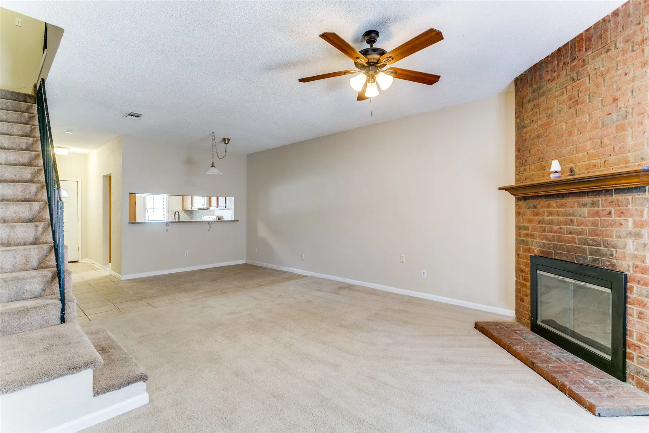 Brick-clad fireplace with a wood mantle, ceiling fan with integrated lighting, and a pass-through window with a breakfast bar