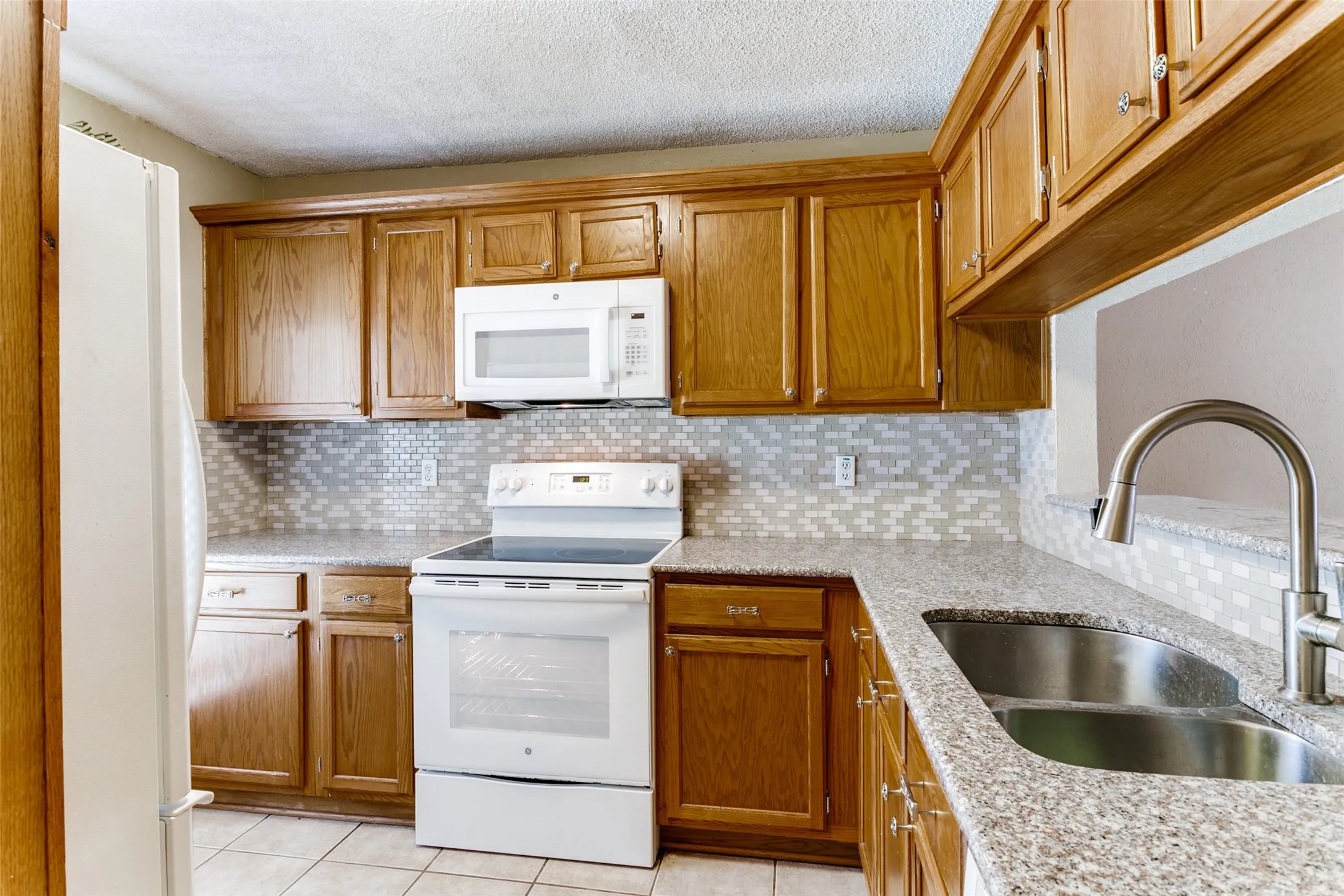 Wood-finish cabinetry, speckled countertop, tile backsplash, stainless steel double basin sink, and an integrated white range
