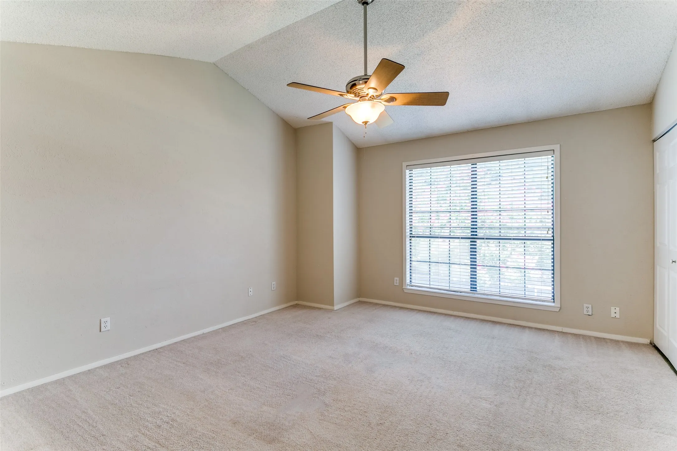 Vaulted ceiling, ceiling fan with integrated lighting, large window with blinds, light-colored carpet flooring, neutral paint palette