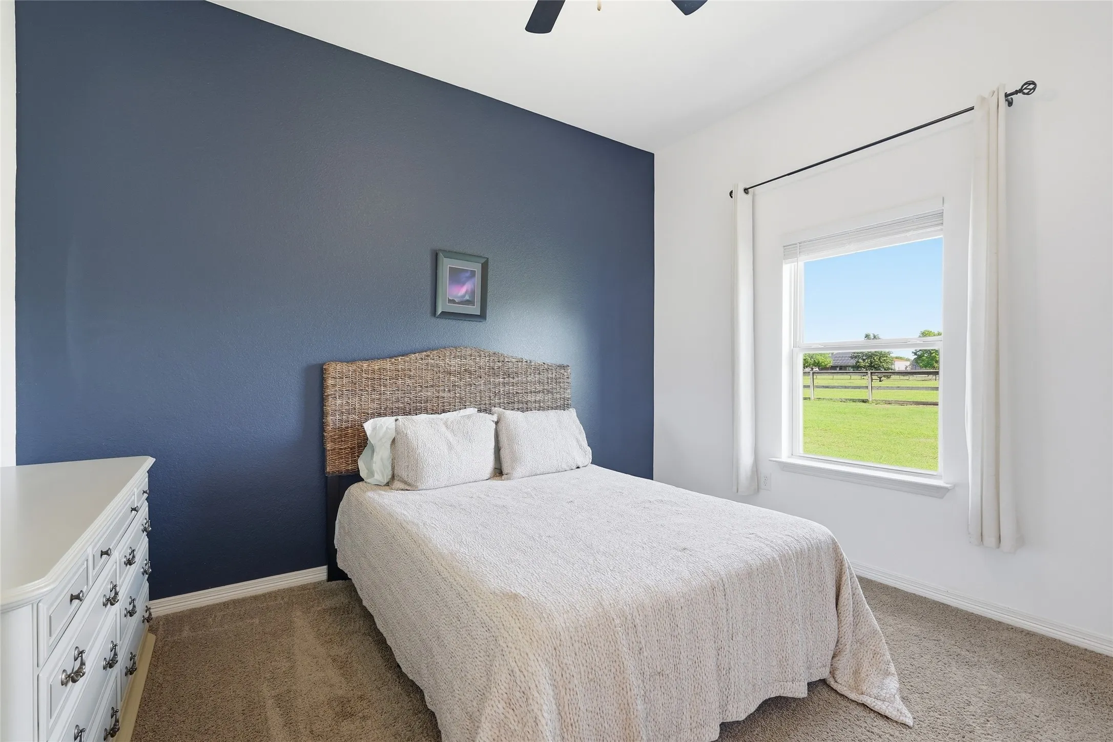 Neutral tone carpeting, a white dresser with ornate hardware, a window with a white frame, and a ceiling fan are present