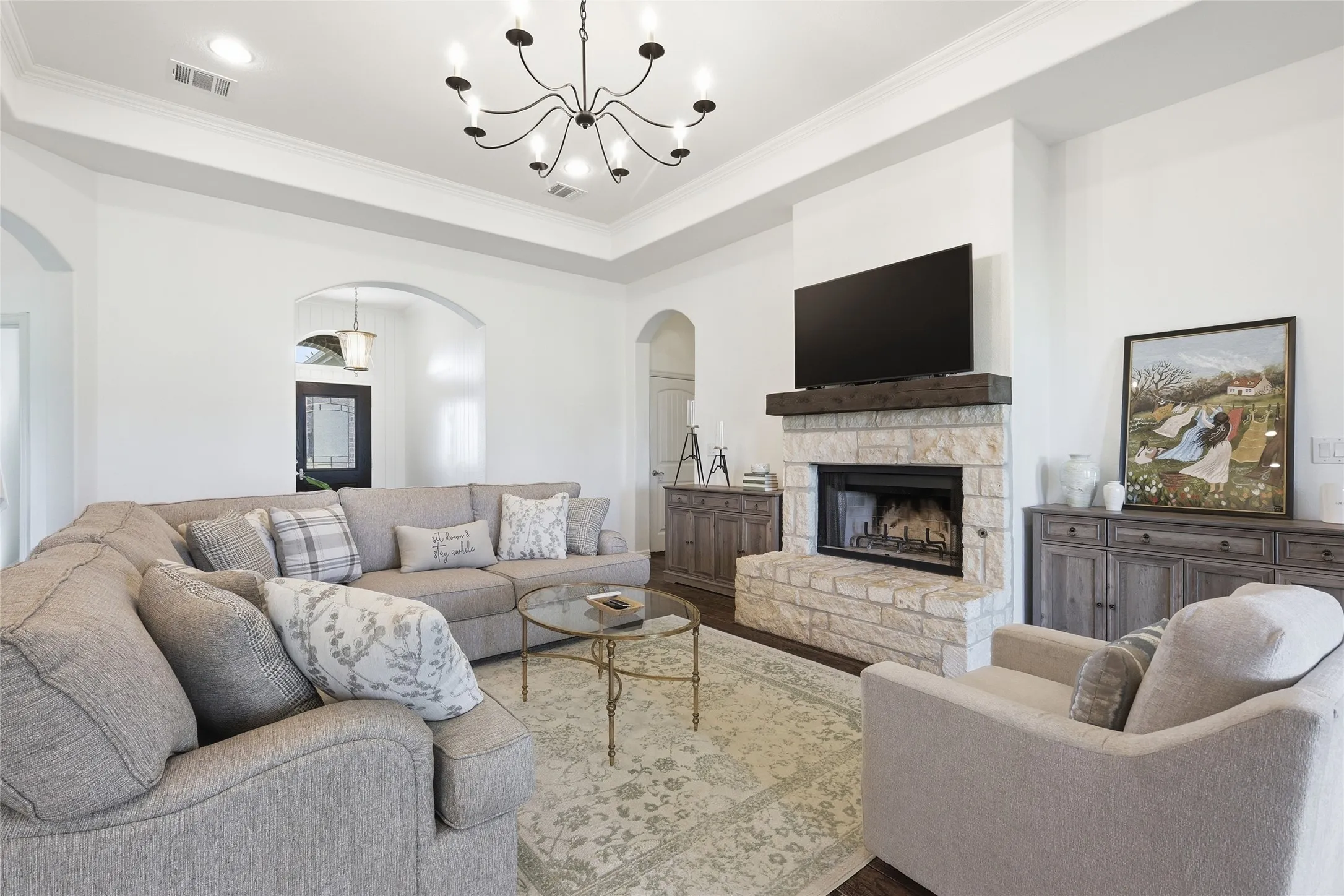 Stone-faced fireplace with a wood mantel, tray ceiling with crown molding, overhead chandelier, arched doorways, and recessed lighting