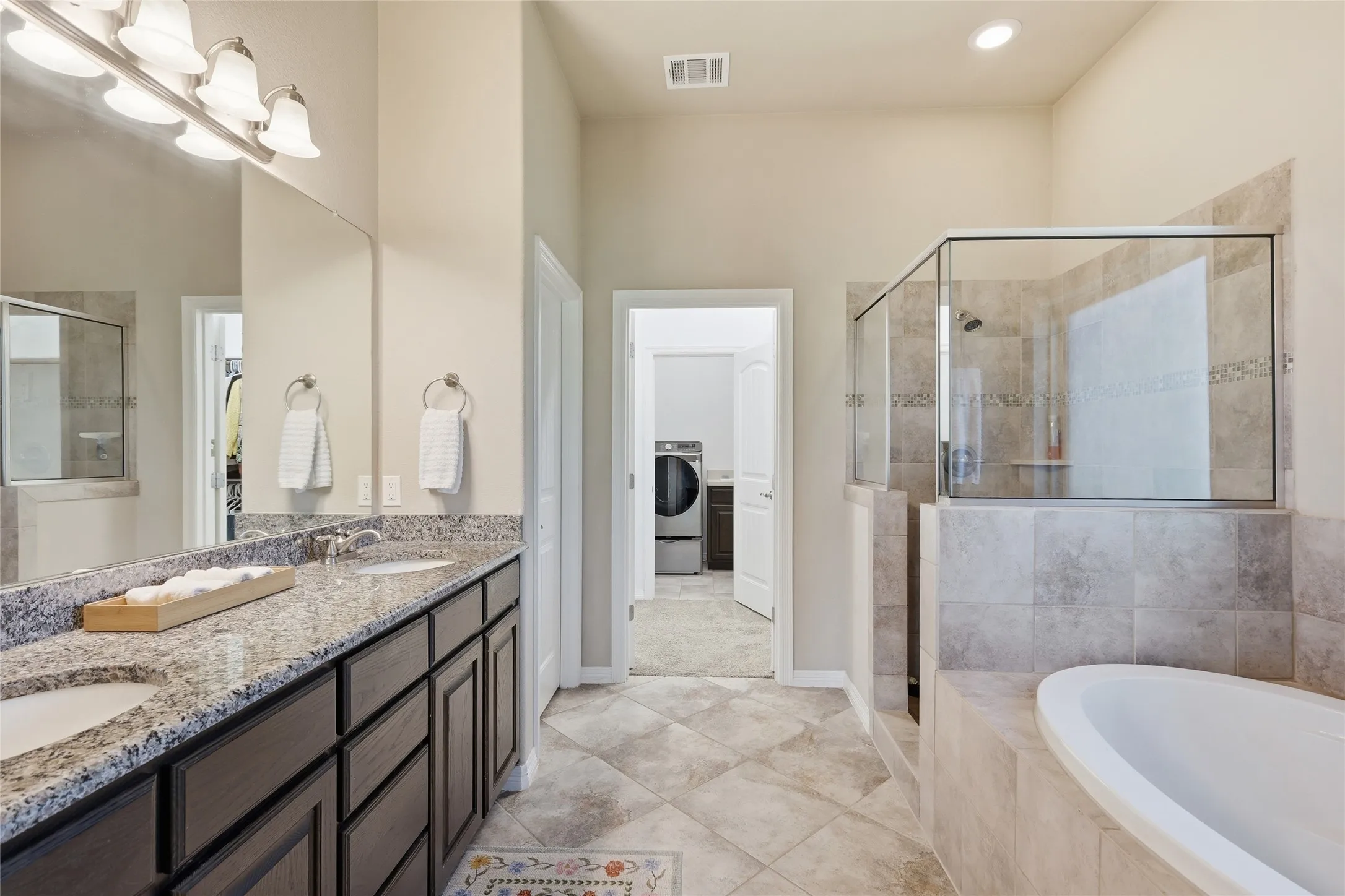 Dual vanity with a granite-style countertop and undermount sinks, a separate glass-enclosed shower, a built-in soaking tub, and tile flooring