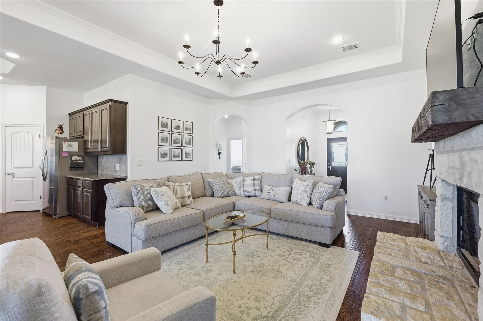 Tray ceiling with chandelier, stone fireplace with wood mantel, wood-finish flooring, dark wood cabinetry, and arched doorways