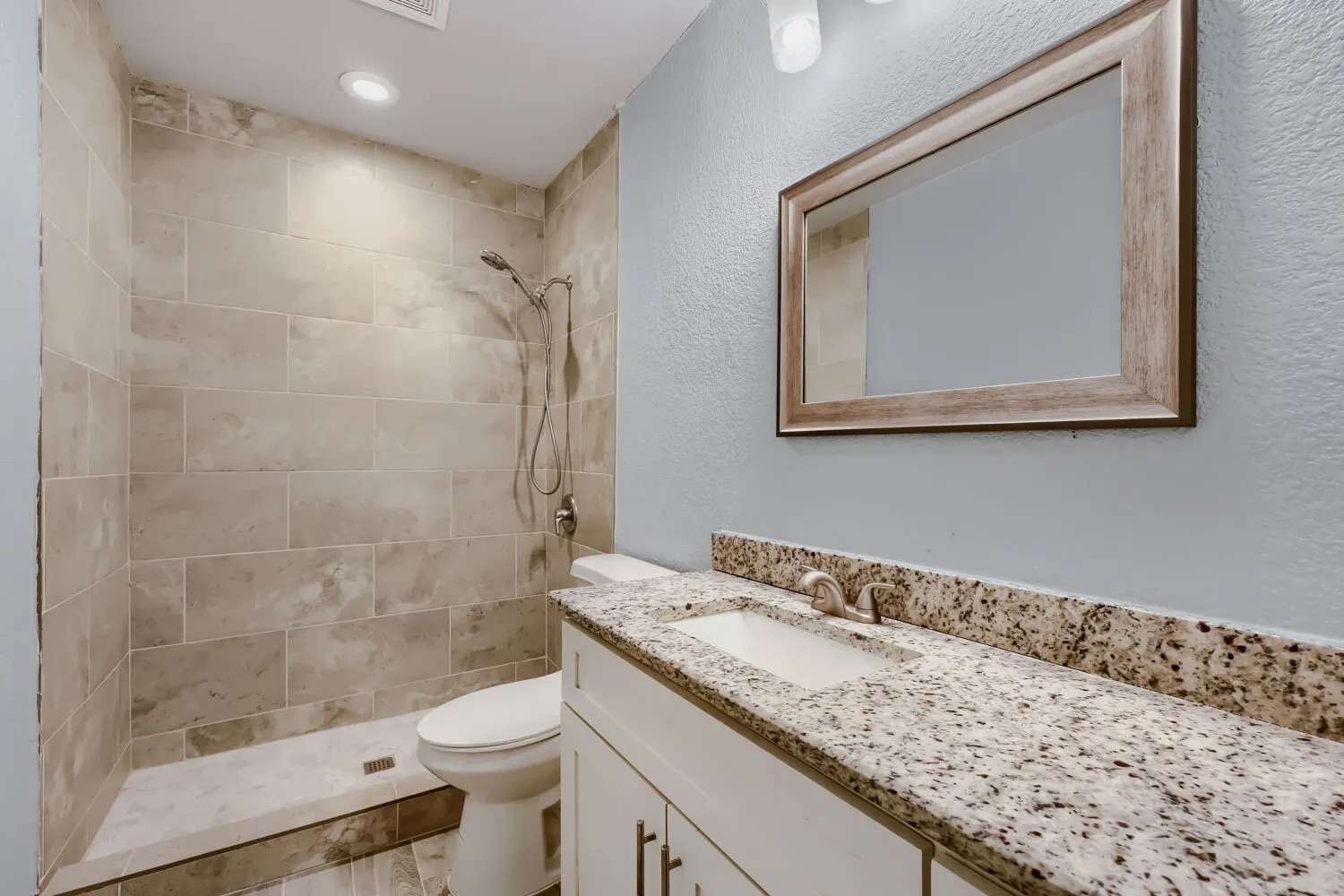 Walk-in shower with large format tile, built-in recessed lighting, white vanity cabinet, granite countertop with integrated sink, and framed mirror