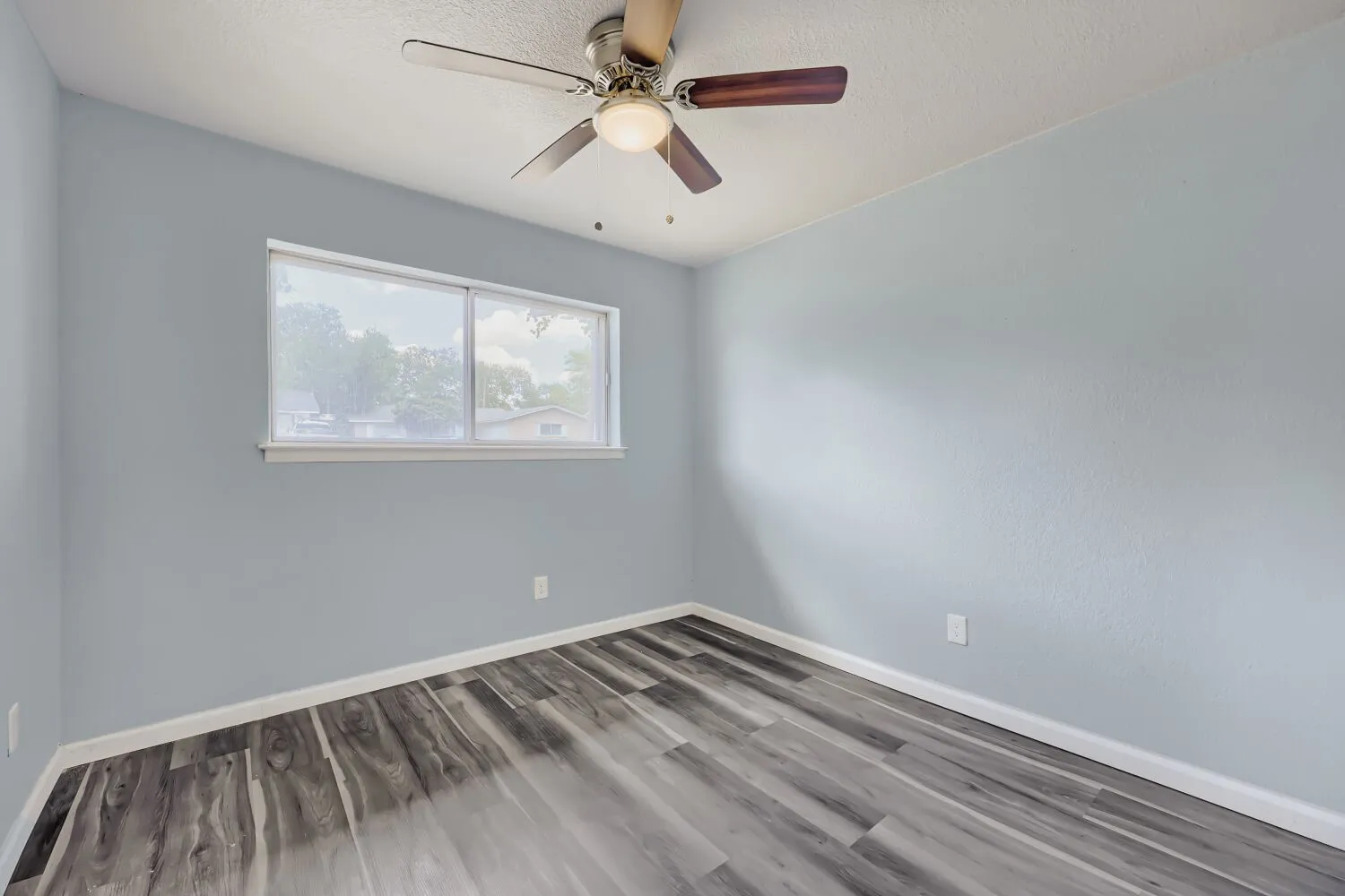 Spacious room featuring wood-finish flooring, light blue walls, a ceiling fan with integrated lighting, and a large window