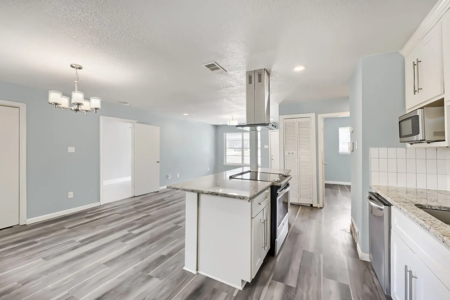 Open-concept living area featuring wood-finish flooring, a kitchen island with cooktop, stainless steel appliances, white cabinetry, and granite countertops
