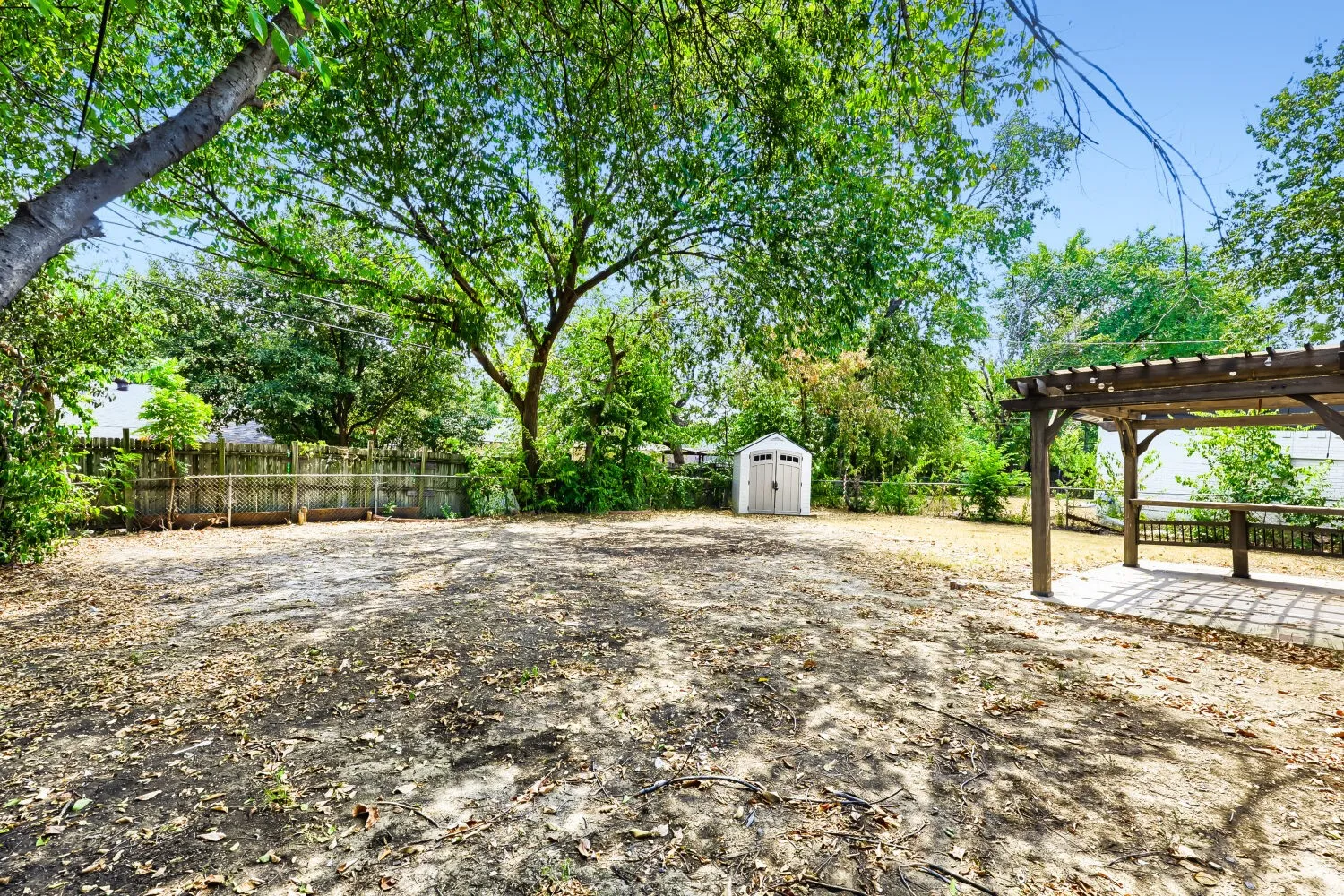 Expansive outdoor area featuring mature trees, a wooden pergola with a concrete patio, a storage shed, and perimeter fencing