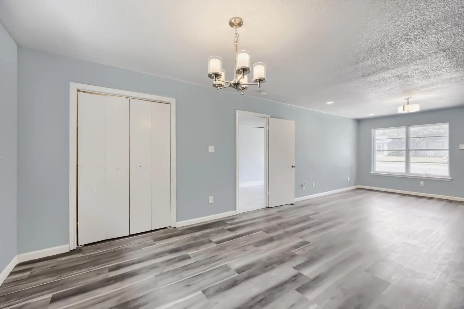 Wood-finish flooring throughout, light blue wall paint, double-pane window, flush-mount ceiling light, and a chandelier with frosted glass shades