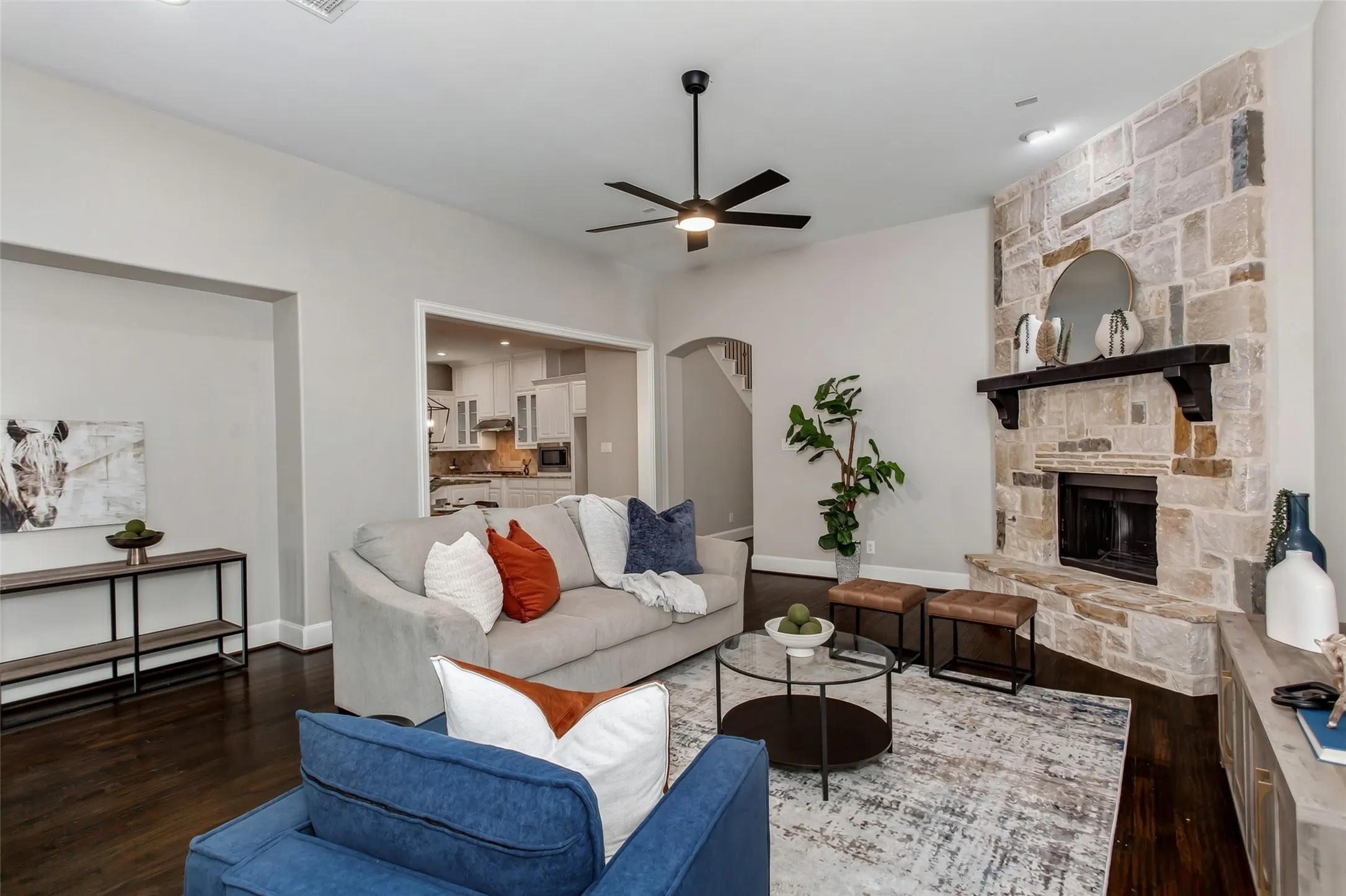Stone-clad fireplace with dark wood mantel, dark wood-finish flooring, ceiling fan with integrated lighting, and arched entryways