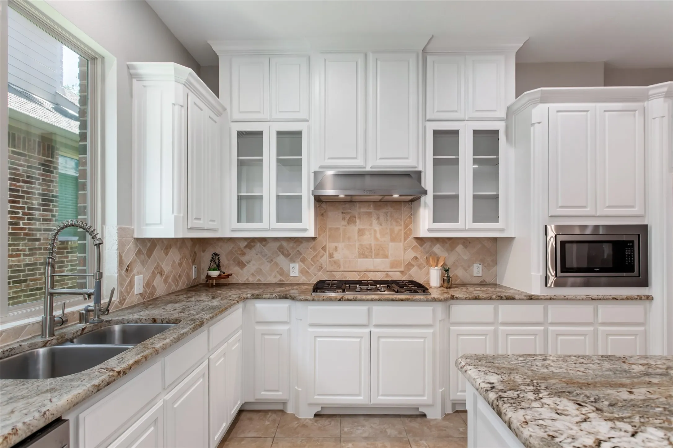 Gas cooktop with a stainless vent hood set against a tumbled stone backsplash with herringbone inlay.
Glass-front upper cabinets, built-in microwave, walk-in pantry door, and island seating.