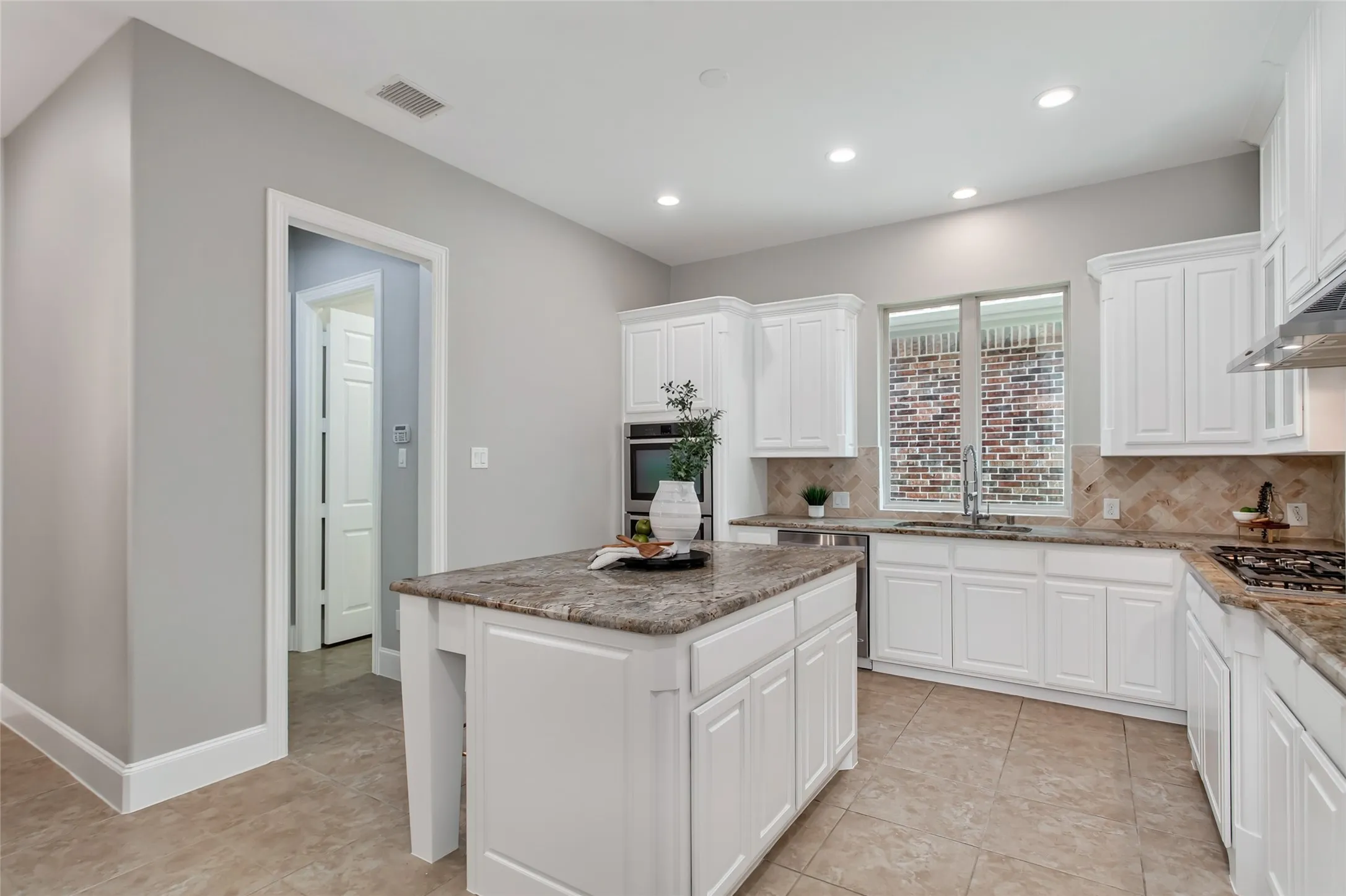 Undermount sink with a pull-down faucet set below a window, flanked by white raised-panel cabinetry
and granite counters. Stainless dishwasher, double wall ovens, and a doorway to the utility hall.