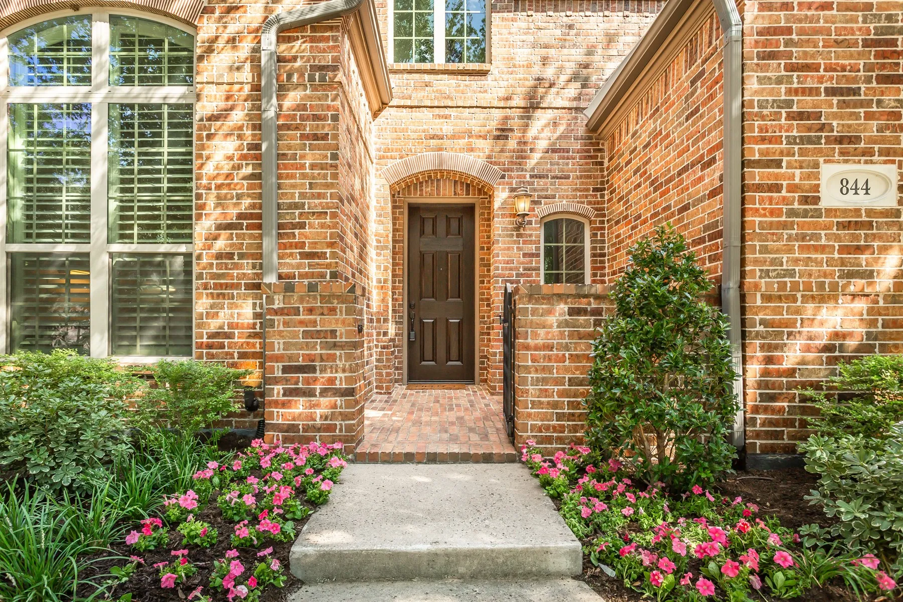 Arched brick entry with a wrought iron gate, brick-paver courtyard, and dark stained front door. Flower
beds with fresh plantings from the 2026 landscape refresh frame the walkway.