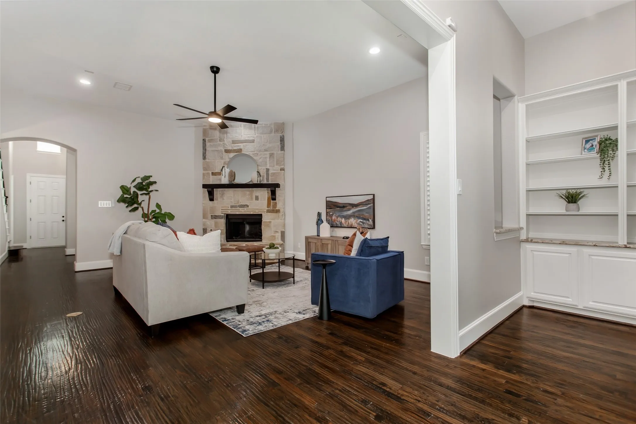Wide view of the stone fireplace, the arched entry to the foyer, and a built-in bookcase with granite
ledge and lower cabinets. Hand-scraped hardwoods and a new 2026 ceiling fan.