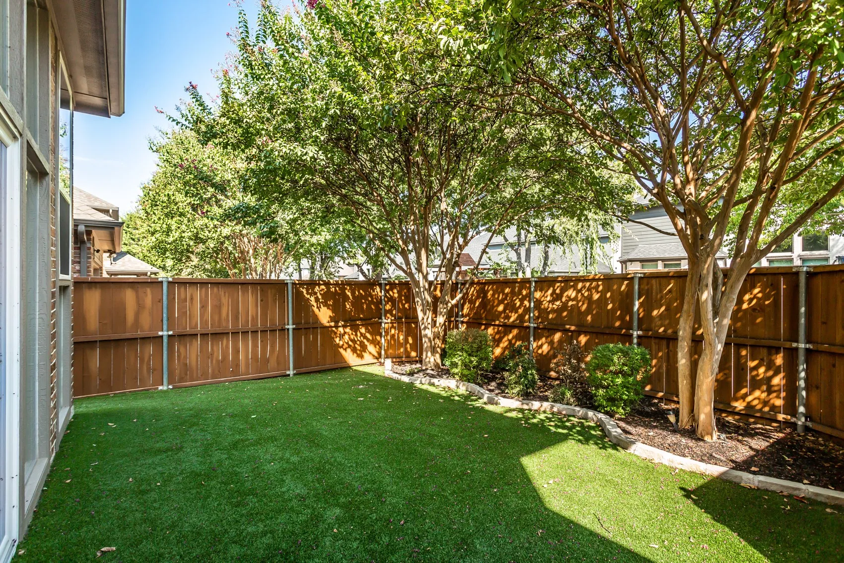 Artificial turf backyard with no mowing required, a stained cedar privacy fence on steel posts, and
mature crape myrtles. Stone-edged planting bed with shrubs along the fence line.