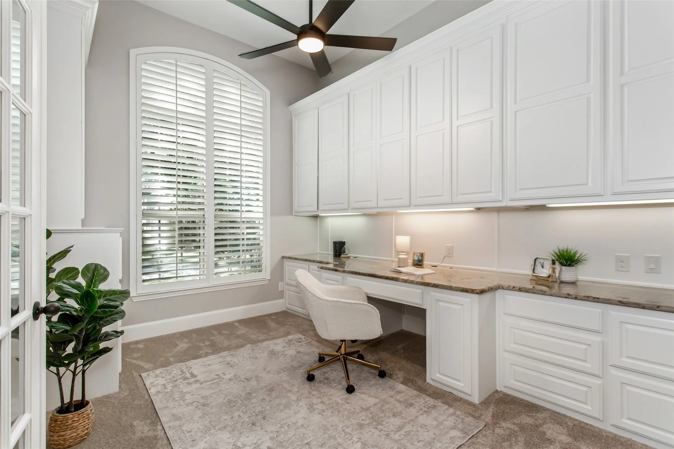 Private study with a wall of built-in cabinetry, a granite desktop with under-cabinet lighting, and an
arched window with plantation shutters. New 2026 carpet and a new ceiling fan with an integrated LED
light.