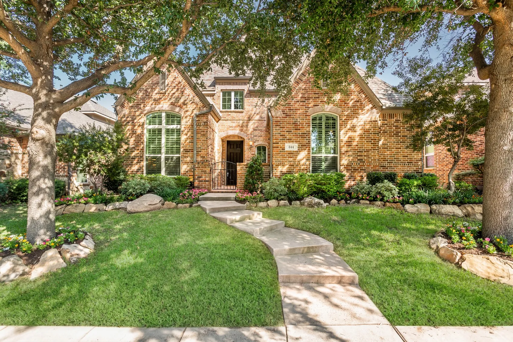 Two-story brick elevation with arched windows, plantation shutters, and a gated front entry under
mature shade trees. A curved walkway leads through landscaping refreshed in 2026 with
boulder-edged beds and seasonal color.